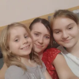 Zdjęcie selfie przedstawia uśmiechniętą kobietę w czerwonej sukience z motywem flamingów, obejmującą dwie dziewczynki, blondynkę w szarej koszulce w serduszka i nastolatkę w białym T-shircie,...