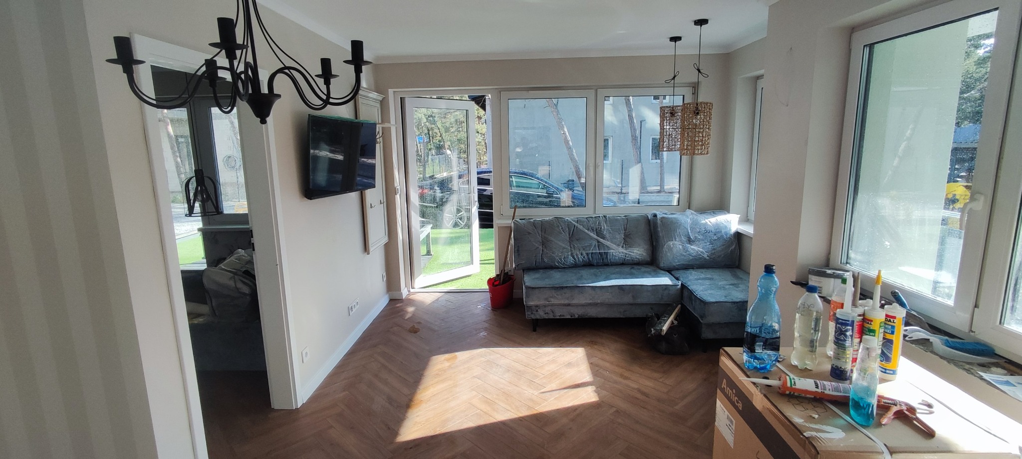 Salon w trakcie wykańczania: parkiet ułożony w jodełkę, sofa zabezpieczona folią, widoczne materiały budowlane i otwarte drzwi na zewnątrz.