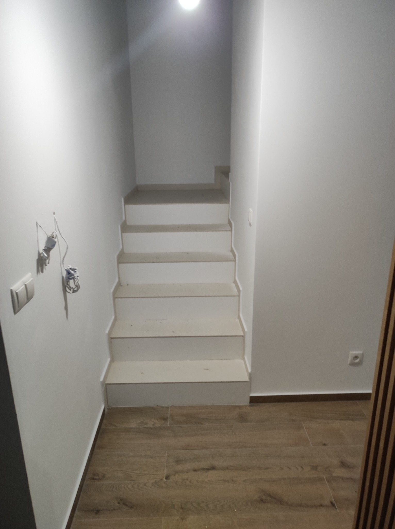 Klatka schodowa w trakcie wykańczania, widoczne białe schody z szarymi krawędziami, drewnopodobna podłoga, białe ściany z niedokończonymi instalacjami elektrycznymi i gniazdkiem.