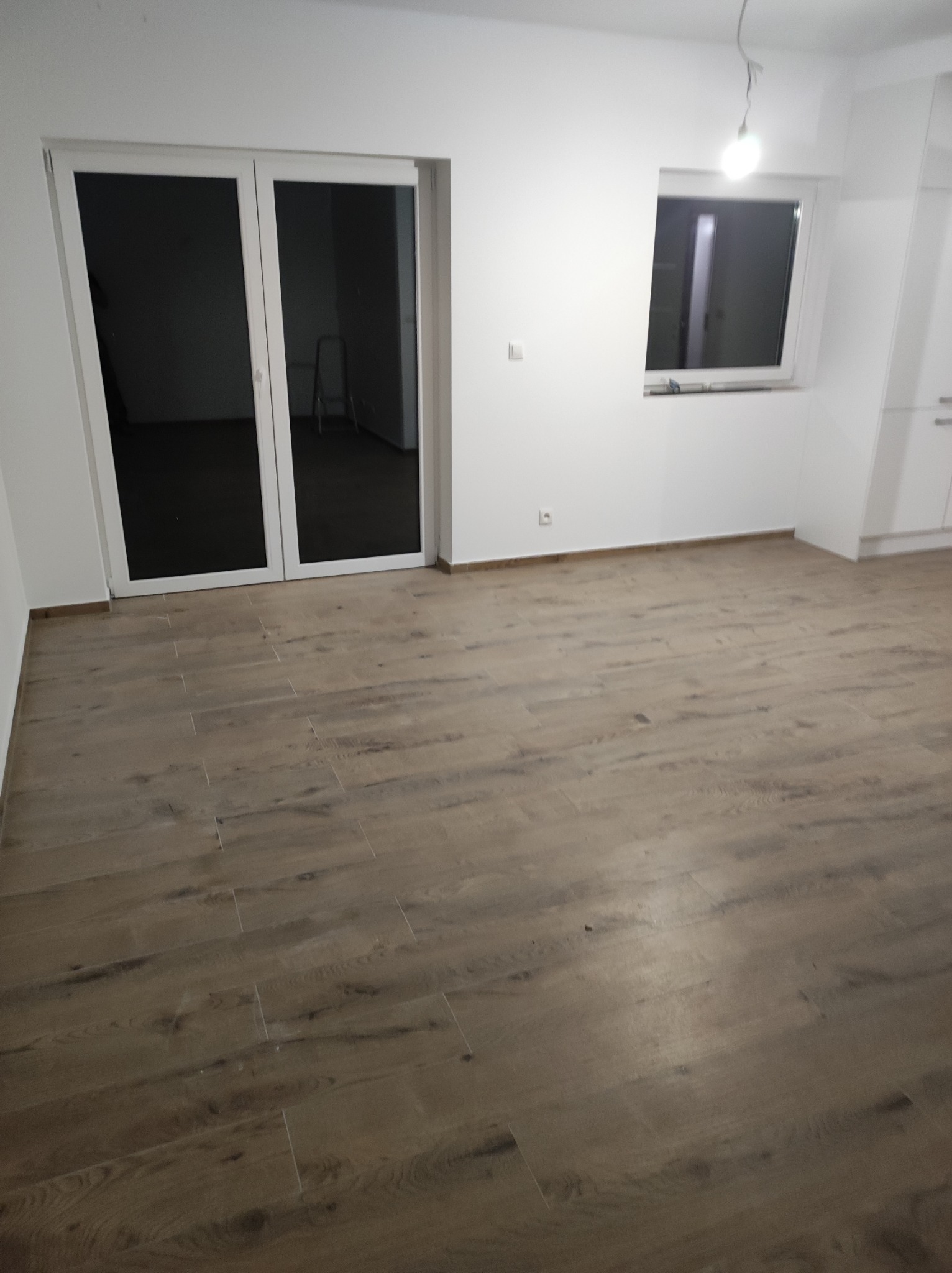 Puste, jasne pomieszczenie z drewnopodobną podłogą z płytek, białymi ścianami, oknem i drzwiami balkonowymi; widoczna drabina w tle za drzwiami.