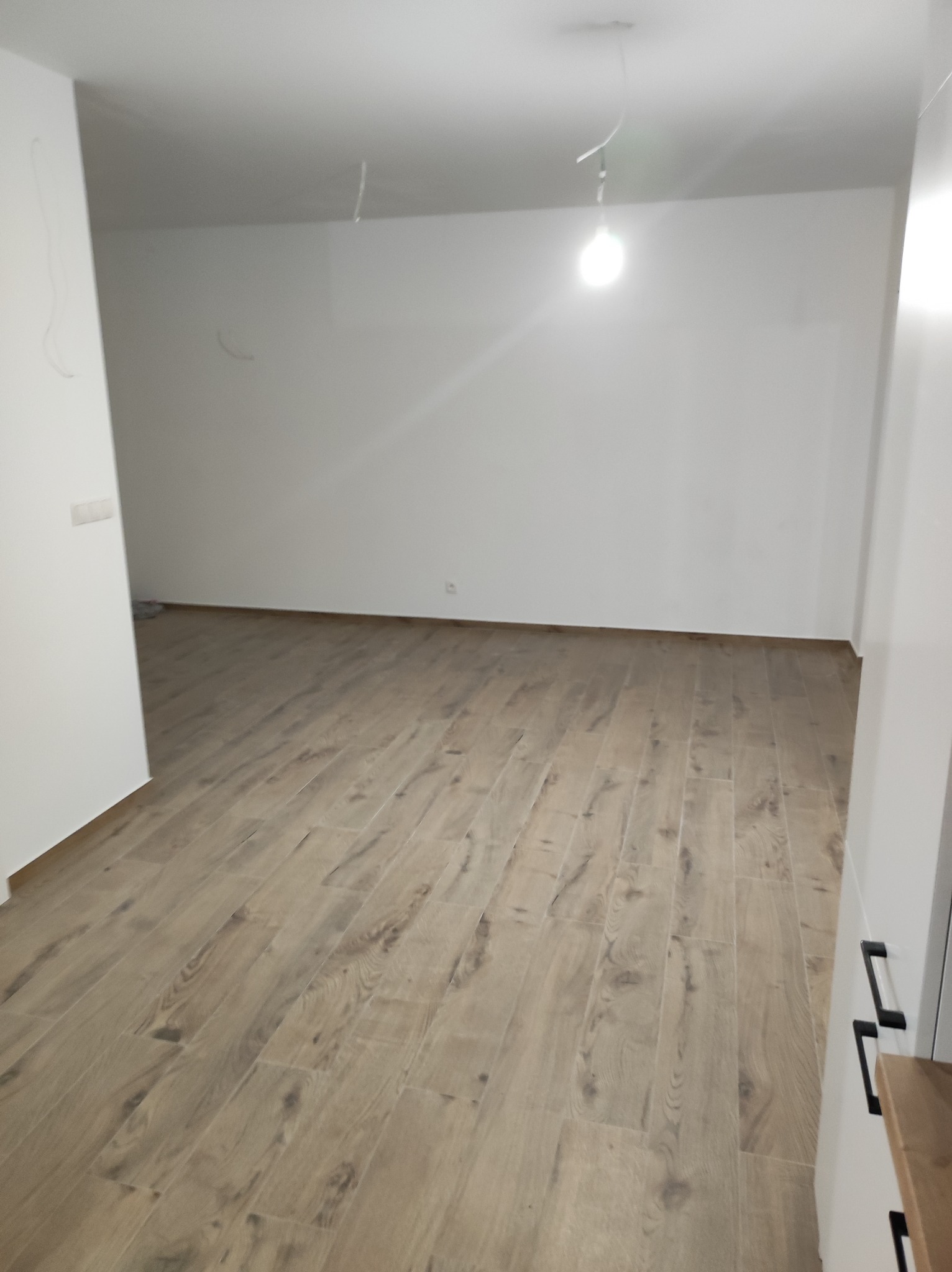 Puste, jasne pomieszczenie po remoncie z drewnopodobną podłogą, widoczne kable zwisające z sufitu i surowa żarówka.