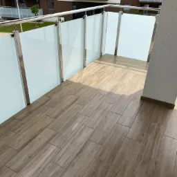 Wykończony balkon w nowoczesnym stylu z podłogą z płytek imitujących drewno, szklane balustrady z metalowymi ramami i widokiem na budynek mieszkalny.