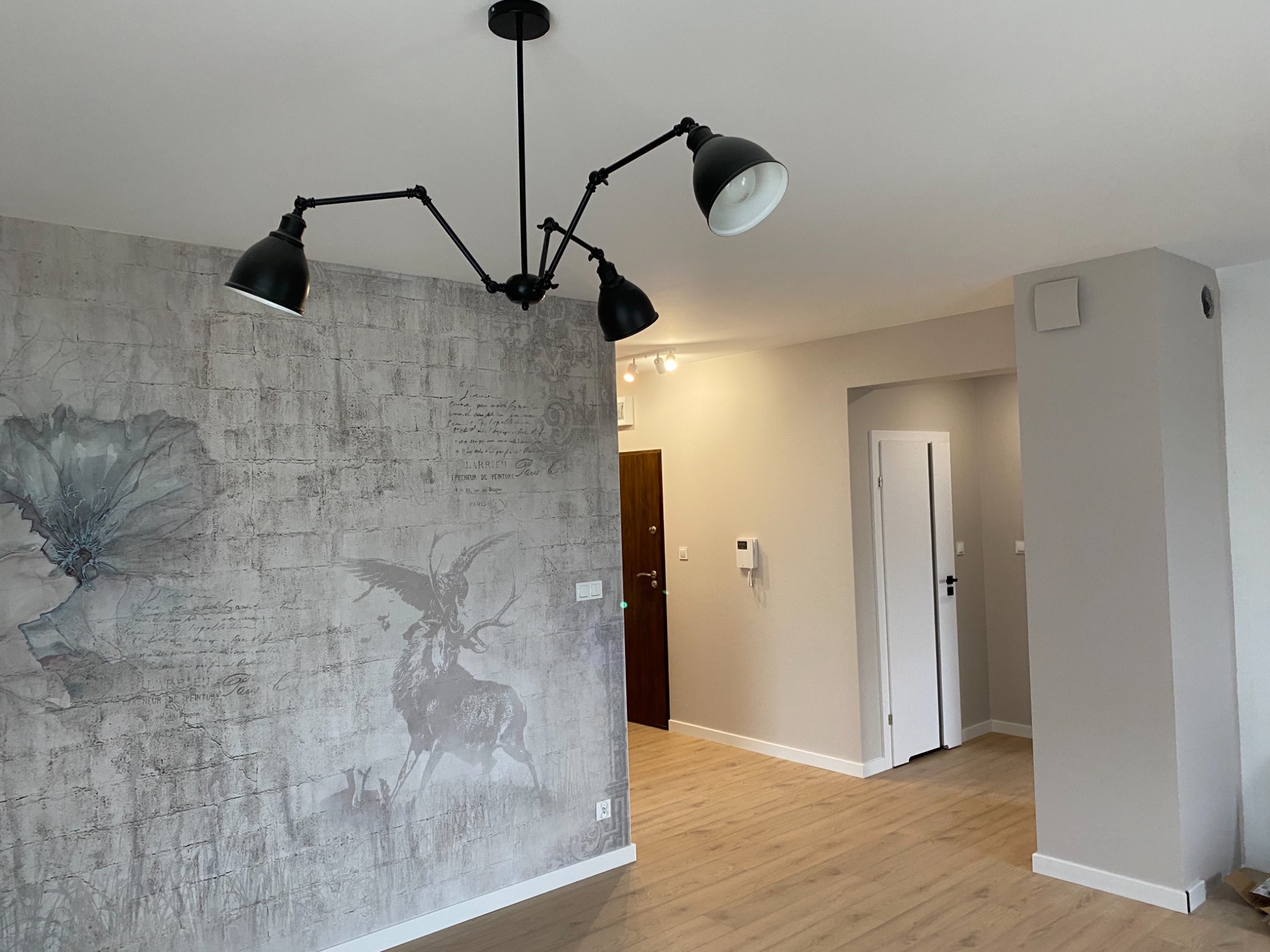 Pokój po odświeżeniu z szarą tapetą w stylu vintage z motywem roślinnym i zwierzęcym, drewnianą podłogą, nowoczesną czarną lampą sufitową oraz świeżo pomalowanymi ścianami w odcieniach beżu.