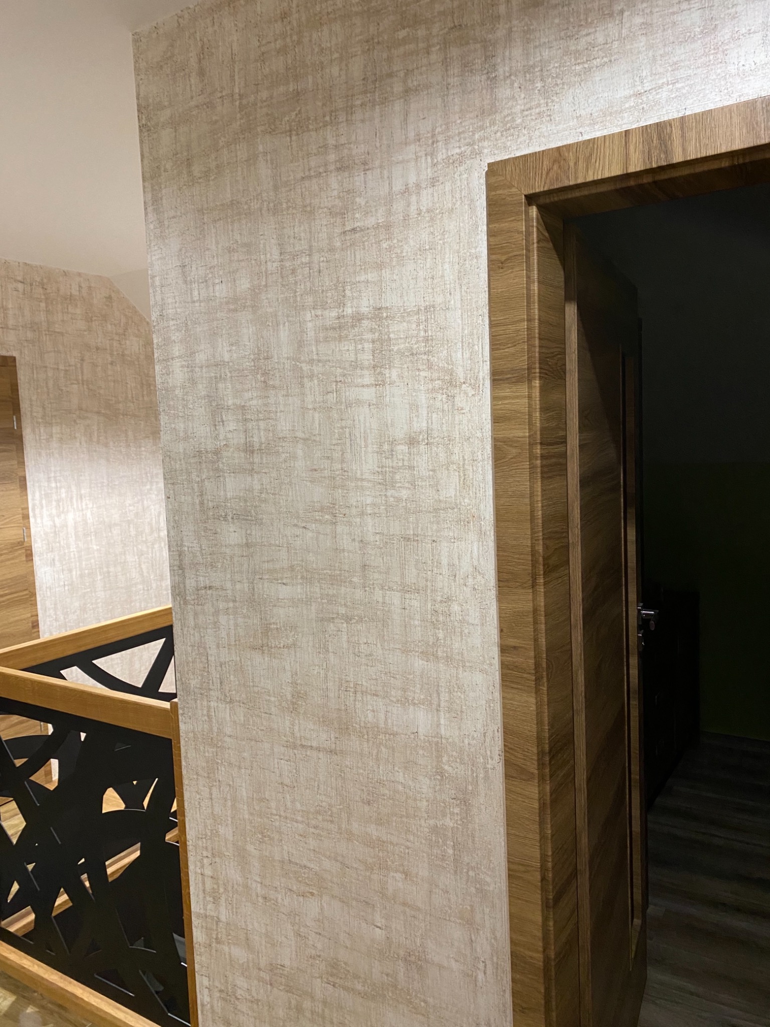 Ściana w odcieniach beżu z delikatną teksturą, sąsiadująca z drewnianą futryną drzwi i czarną, geometryczną balustradą schodów.