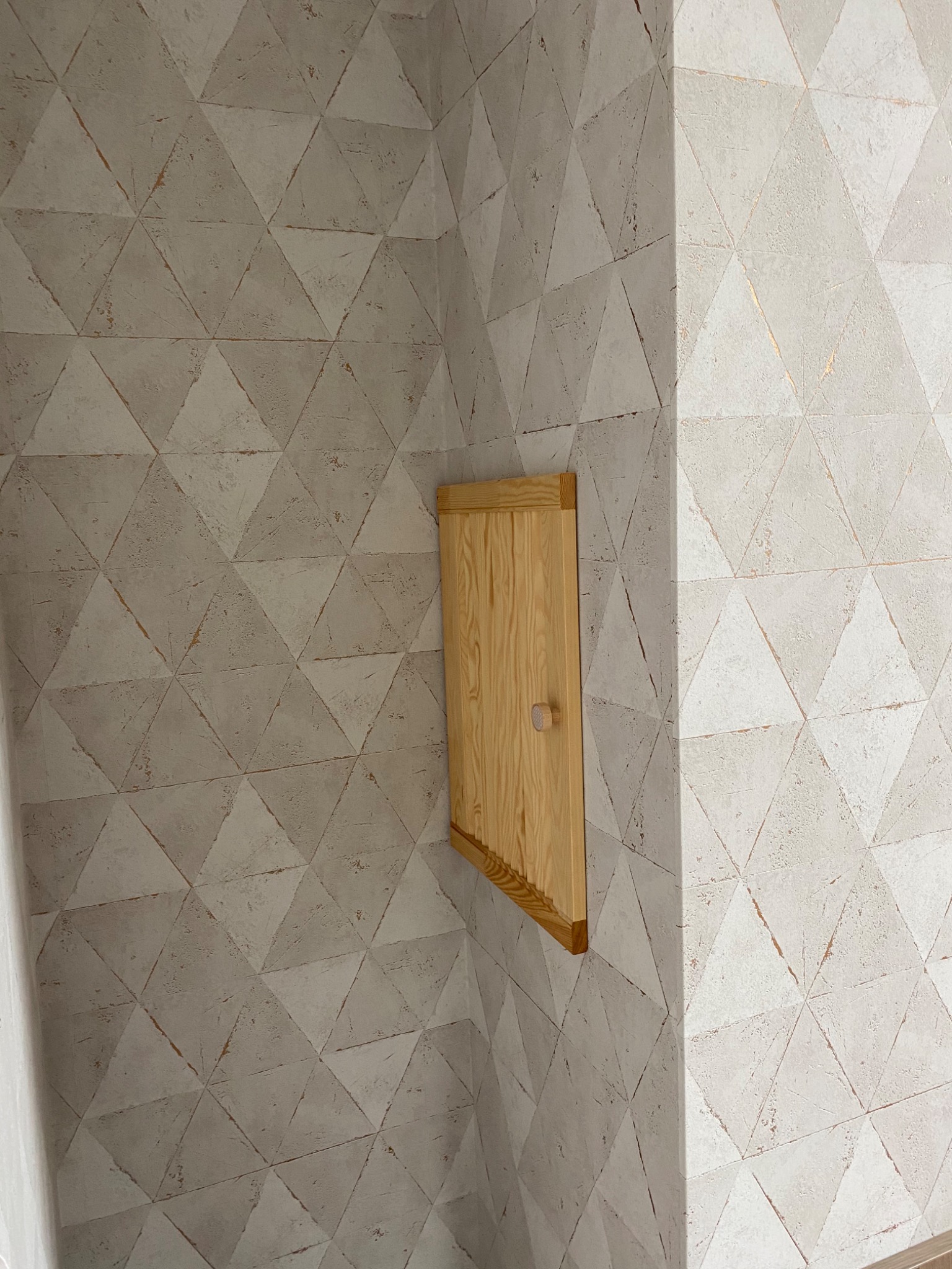 Ściana wykończona w geometryczny wzór trójkątów w odcieniach szarości z drewnianą klapą rewizyjną z okrągłym uchwytem.