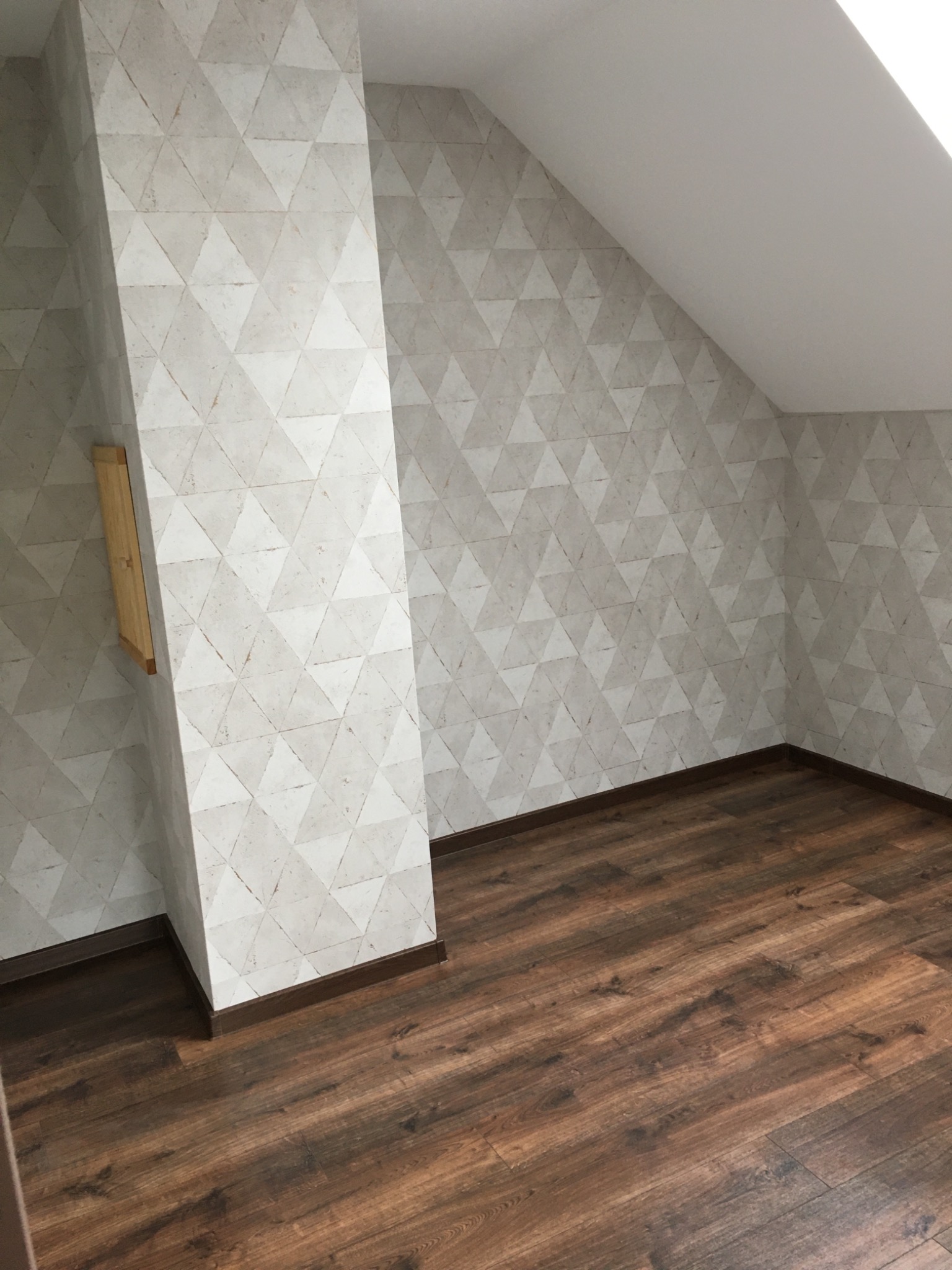 Poddasze po remoncie z drewnianą podłogą i ścianami pokrytymi tapetą w geometryczne wzory, widoczny pionowy słup z ukrytą szafką.