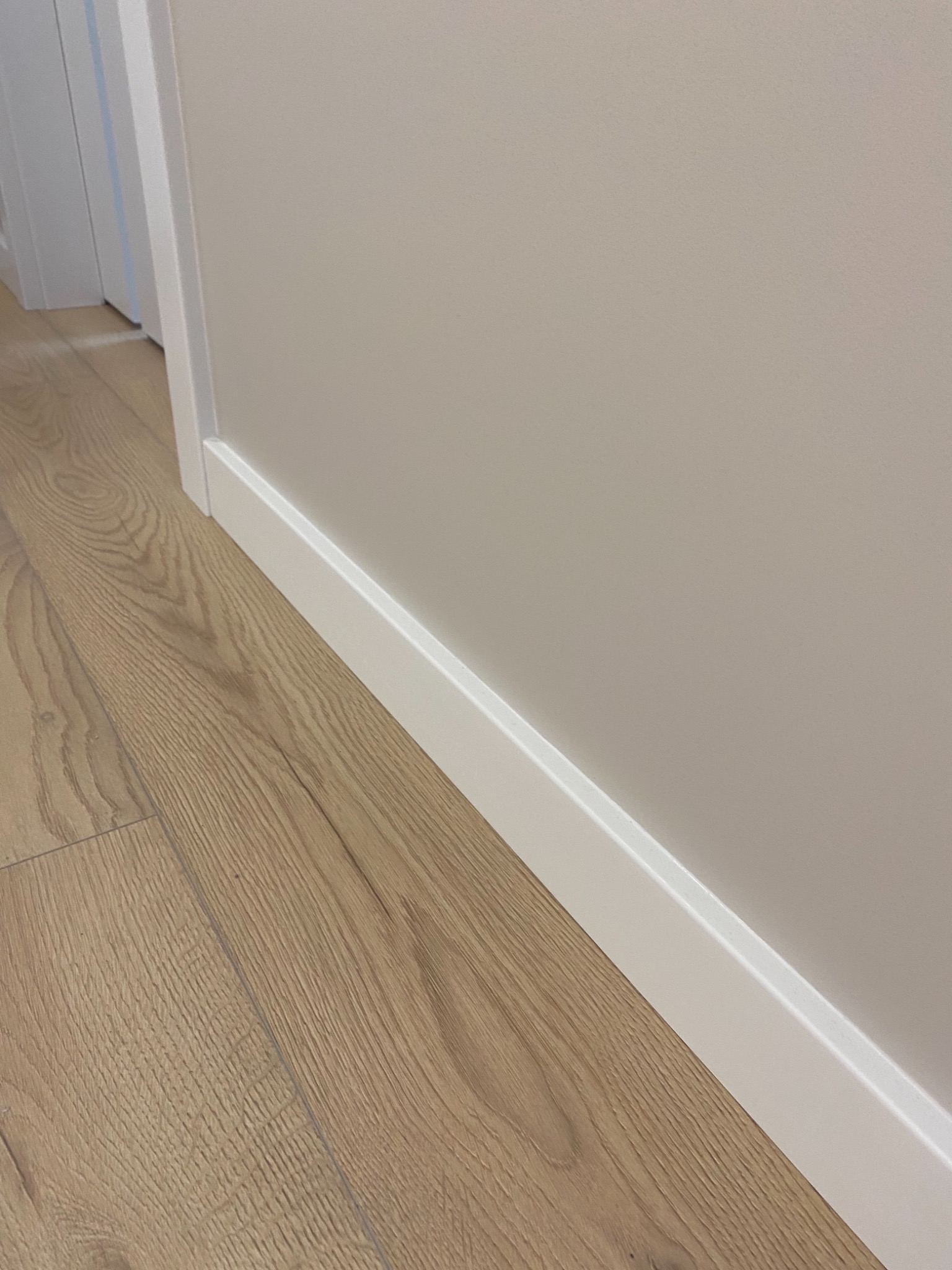 Fragment wnętrza z jasną ścianą, drewnopodobną podłogą i białą listwą przypodłogową oraz obramowaniem drzwi.