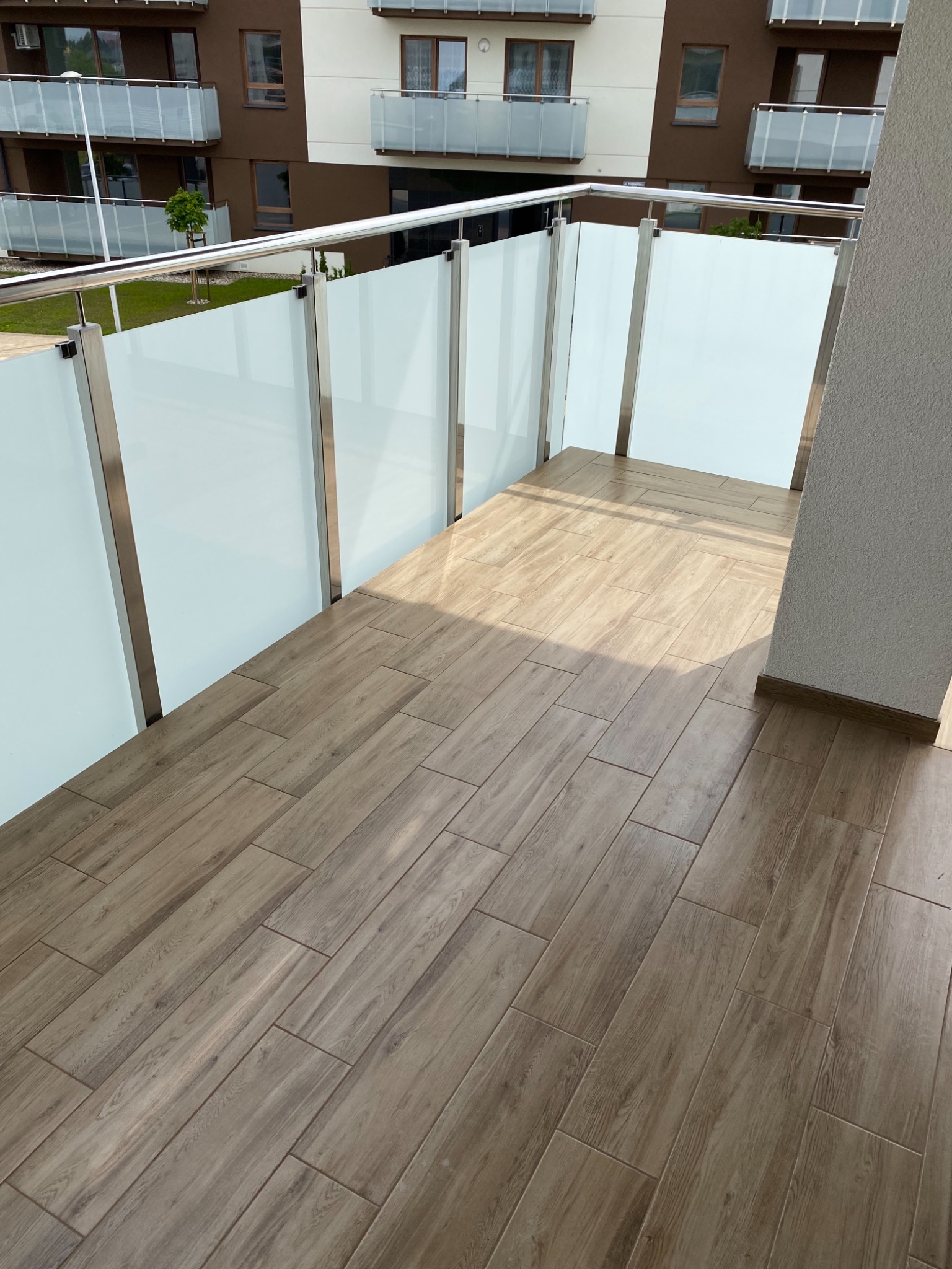 Wykończony balkon w nowoczesnym stylu z podłogą z płytek imitujących drewno, szklane balustrady z metalowymi ramami i widokiem na budynek mieszkalny.