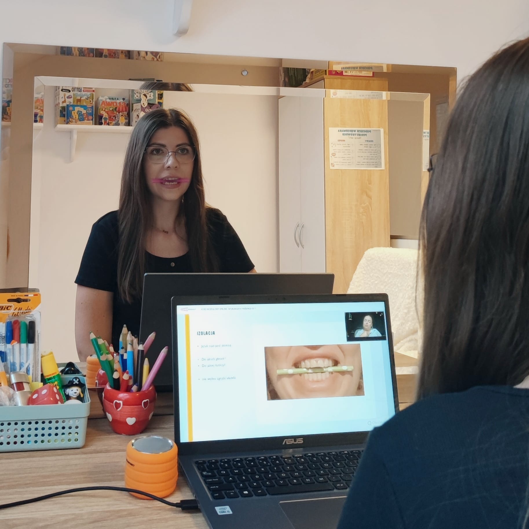 Kobieta z ołówkiem w ustach, odbicie w lustrze, laptop z ekranem pokazującym ćwiczenia artykulacyjne, kolorowe kredki i zabawki na biurku, sesja online.
