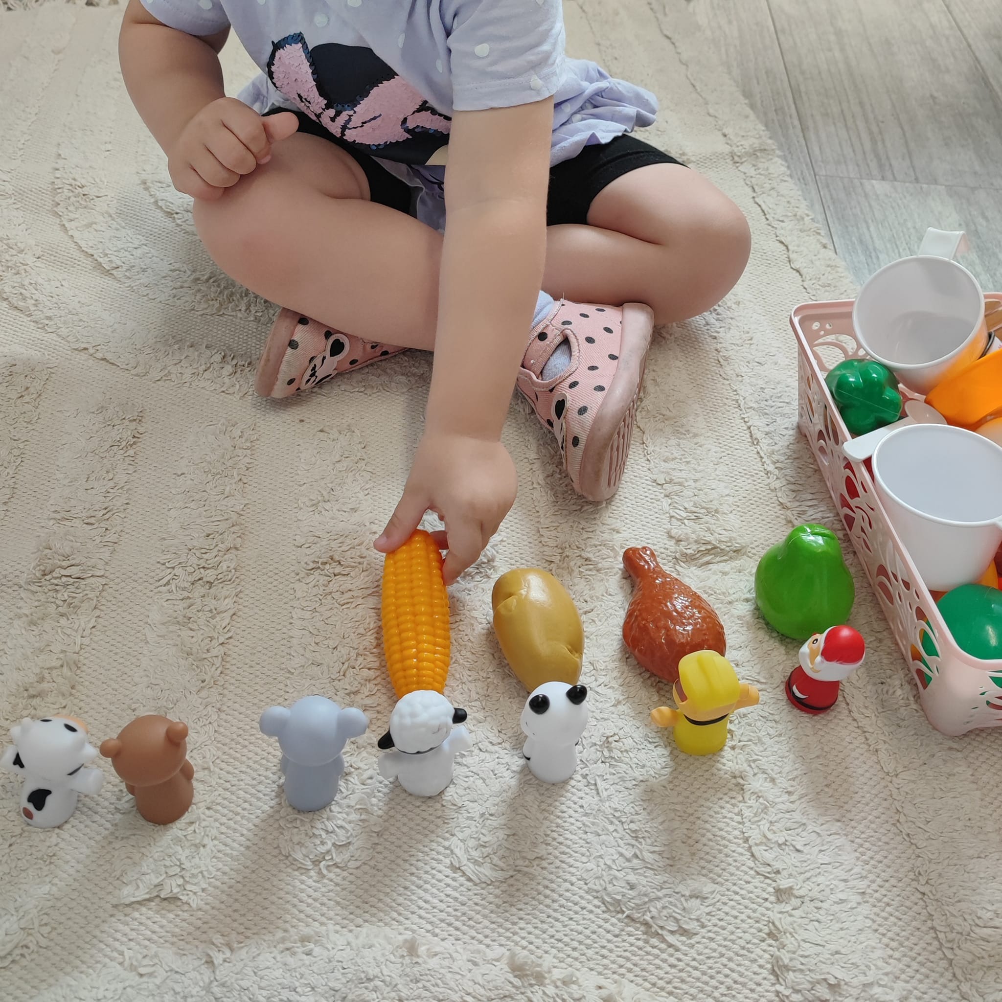 Dziecko bawi się plastikowymi zabawkami imitującymi jedzenie i zwierzęta na kremowym dywanie z frędzlami.