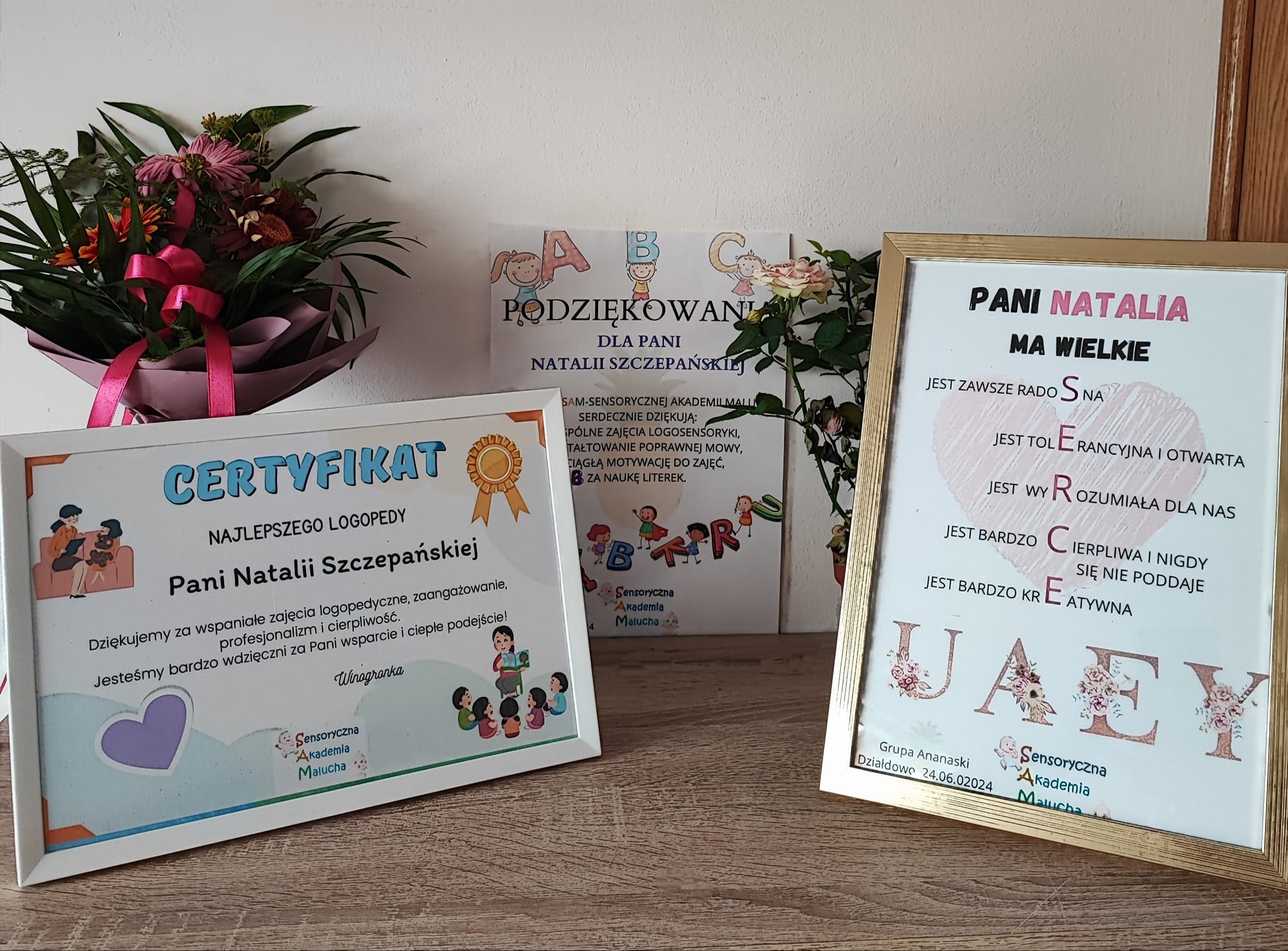 Certyfikat, podziękowanie i ozdobna ramka z dedykacją dla Pani Natalii, otoczone kwiatami, stoją na drewnianym blacie.
