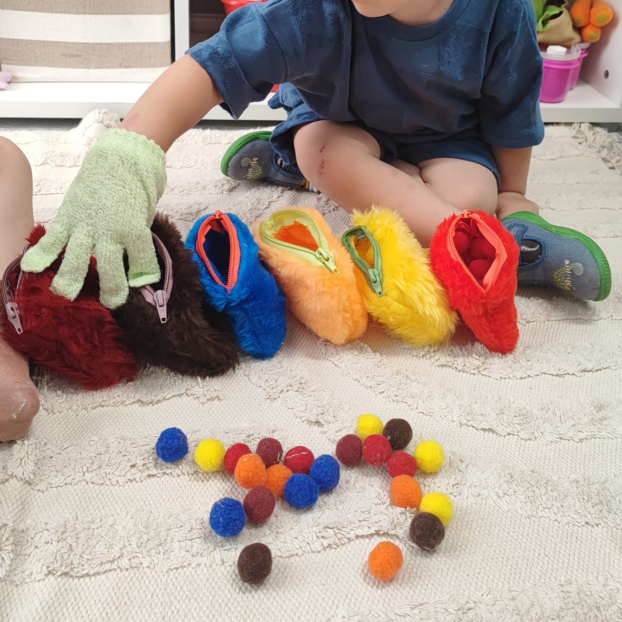 Dziecko siedzi na dywanie bawiąc się kolorowymi, futrzanymi portfelikami z zamkami błyskawicznymi i pomponami
