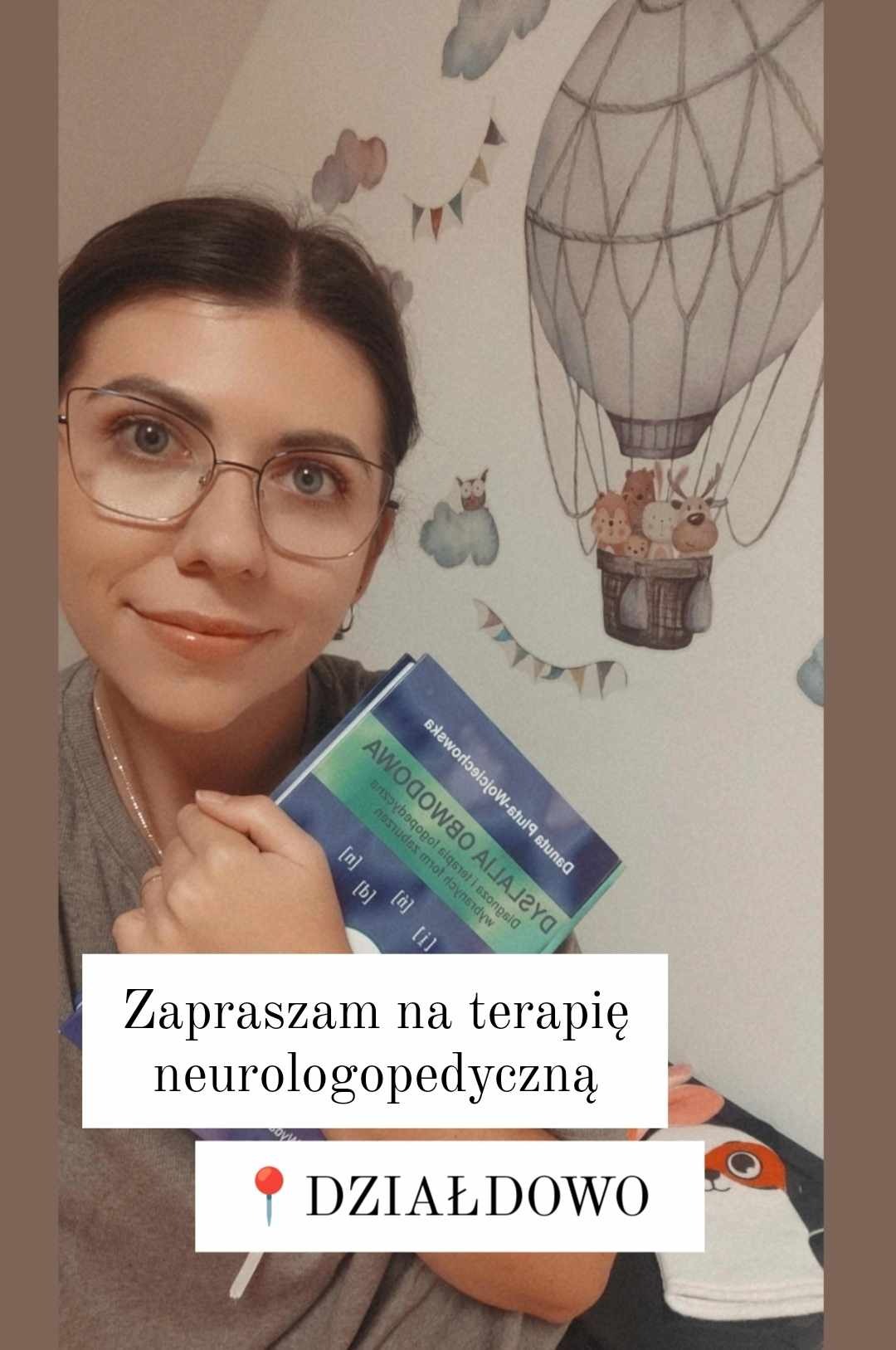 Uśmiechnięta logopedka w okularach trzyma książkę o neurologopedii na tle ściany z naklejką balonu na ogrzane powietrze z pluszakami.