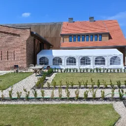 Biały namiot eventowy z oknami na tle ceglanego budynku z drewnianą elewacją i czerwonym dachem, otoczony trawnikiem, żwirowymi ścieżkami i niskimi krzewami.