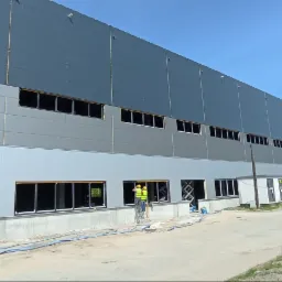 Montaż okien w hali przemysłowej. Dwóch pracowników na podnośniku instaluje okna w szarej hali z panelami. Widoczne otwory okienne i niebieskie węże na ziemi.