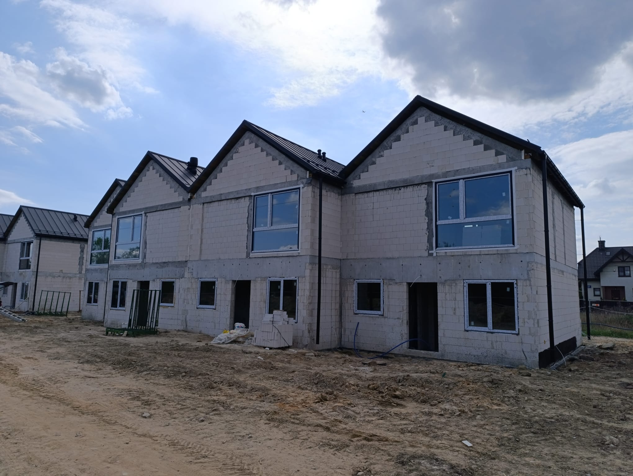 Surowe bryły szeregowców z zamontowanymi oknami, w trakcie budowy, z widocznymi pustakami i elementami instalacji, na tle pochmurnego nieba.