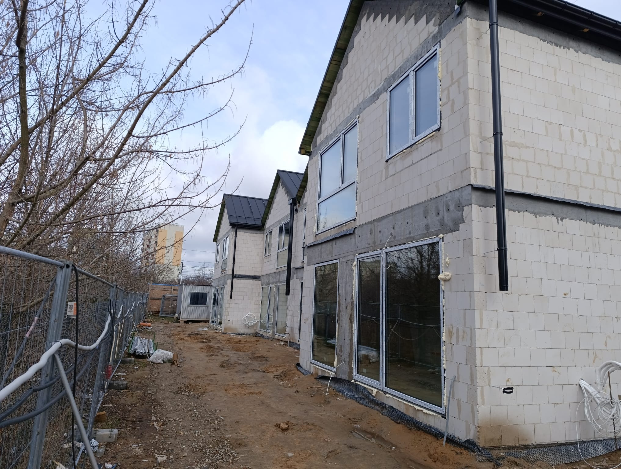 Nowo budowane domy z zamontowanymi oknami PCV. Widoczne surowe ściany z betonu komórkowego i okna z białymi ramami. Teren budowy z ogrodzeniem i ziemią.