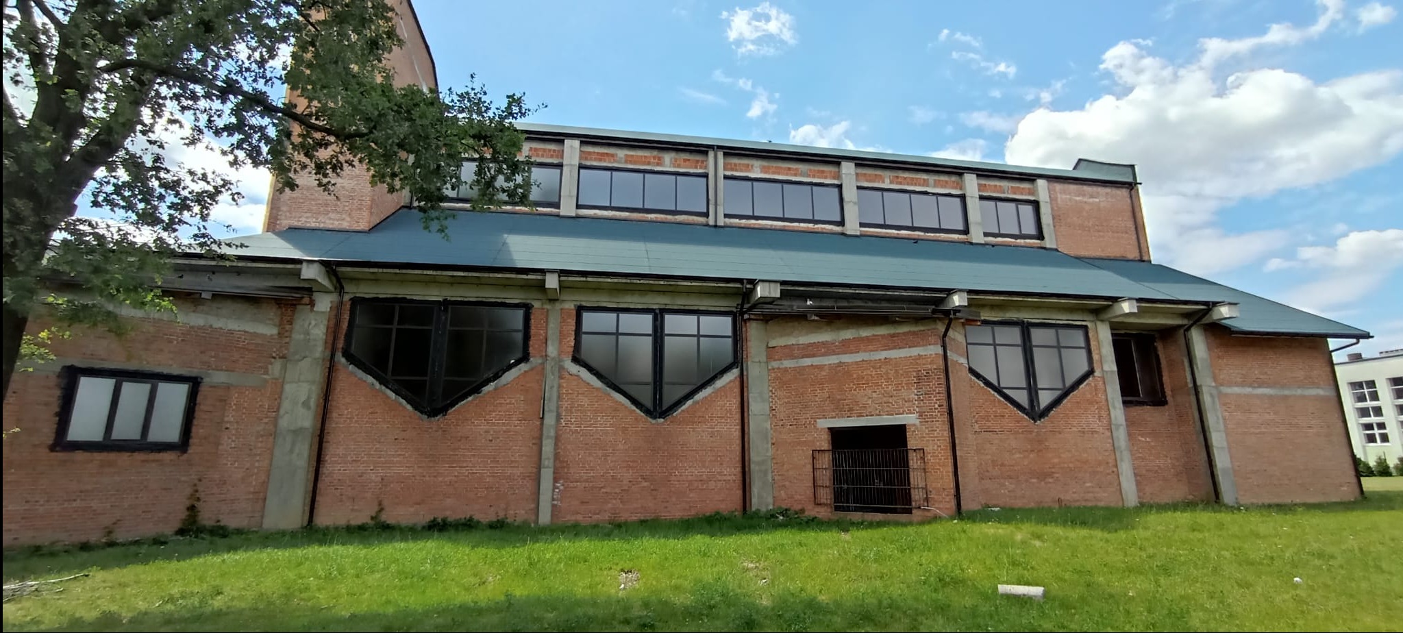 Murowana fasada budynku z nietypowymi, trapezoidalnymi oknami w czarnych ramach, oświetlona naturalnym światłem słonecznym, z fragmentem zieleni w dolnej części kadru.