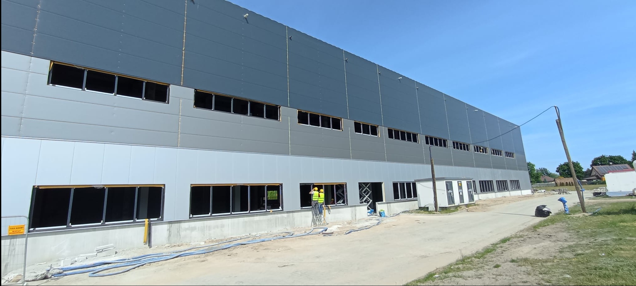 Montaż okien w hali przemysłowej. Dwóch pracowników na podnośniku instaluje okna w szarej hali z panelami. Widoczne otwory okienne i niebieskie węże na ziemi.