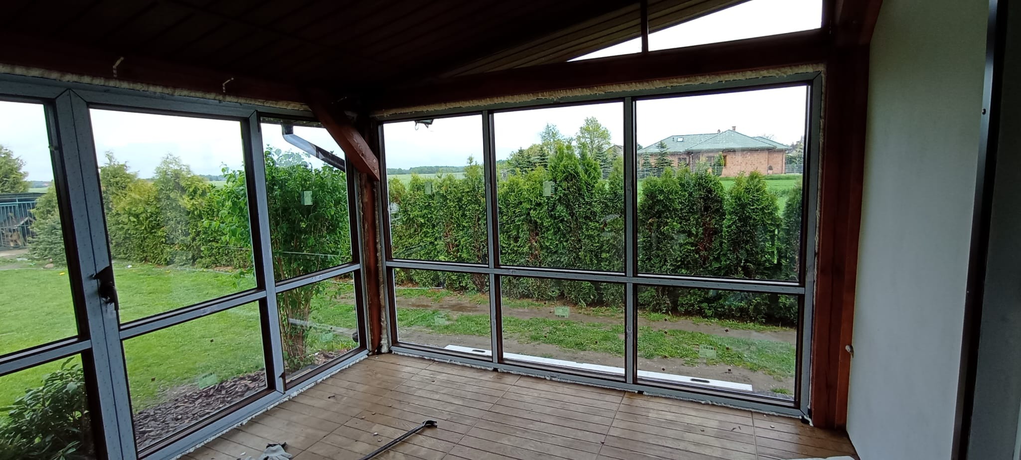 Zadaszony taras z dużymi oknami z widokiem na ogród. Widoczne drewniane elementy konstrukcyjne i uszczelnienia okienne. Na podłodze drewniana podłoga.