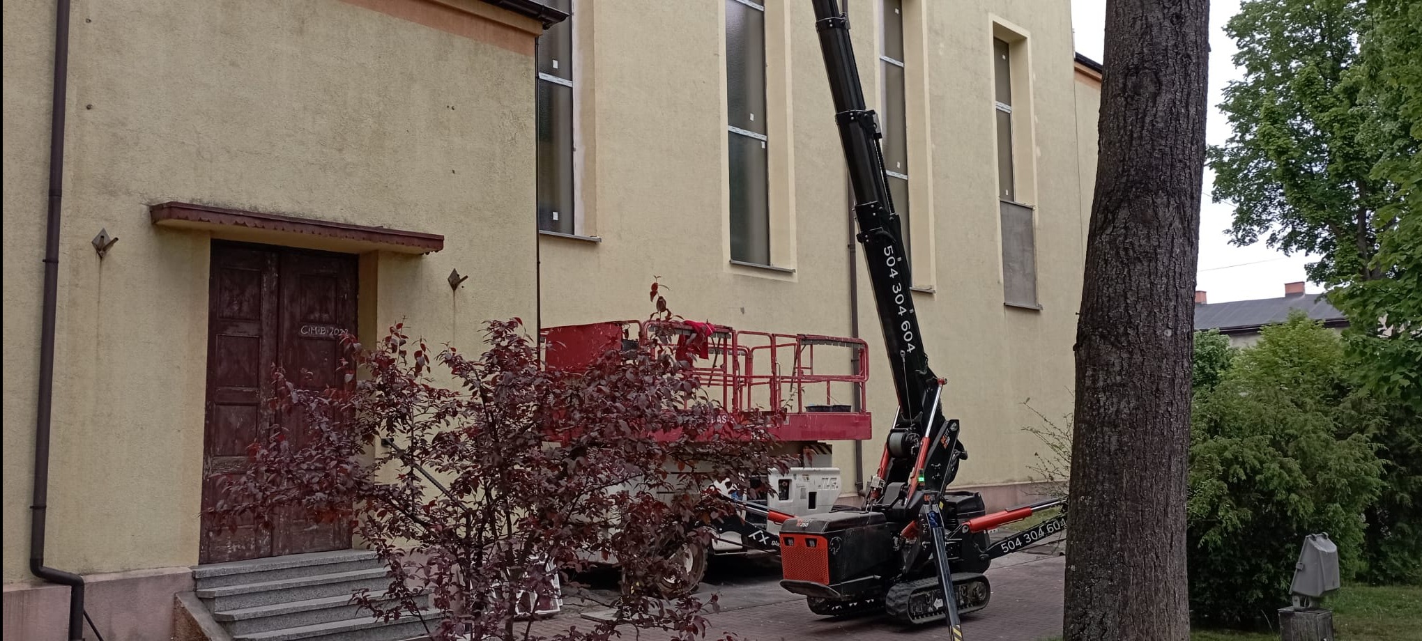 Montaż okien w budynku z wykorzystaniem podnośnika koszowego. Widoczne nowe okna, stary budynek i podnośnik z numerem telefonu firmy.