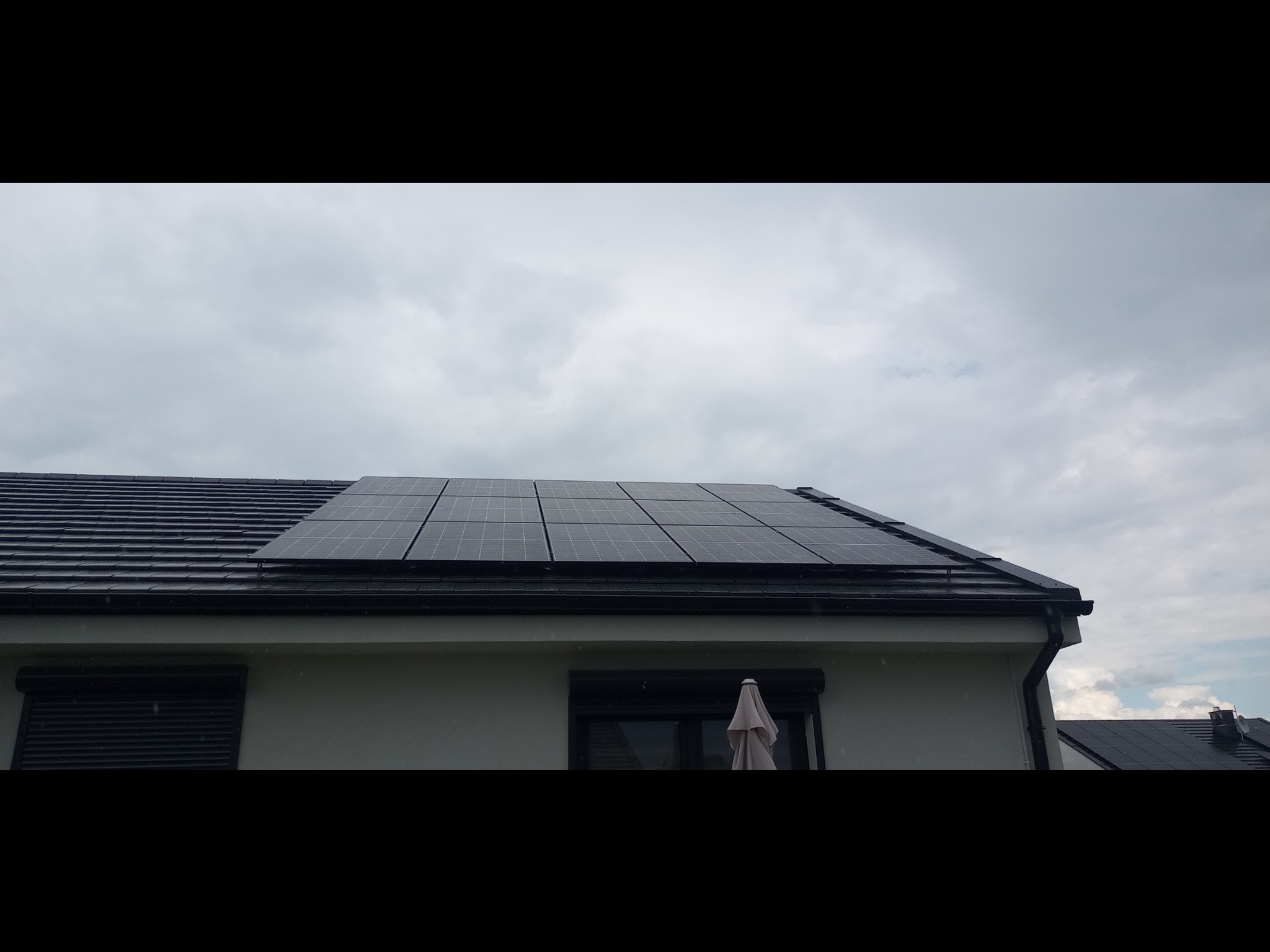 Panele fotowoltaiczne zamontowane na dachu domu jednorodzinnego z ciemną dachówką, widoczne okna z roletami zewnętrznymi i pochmurne niebo.