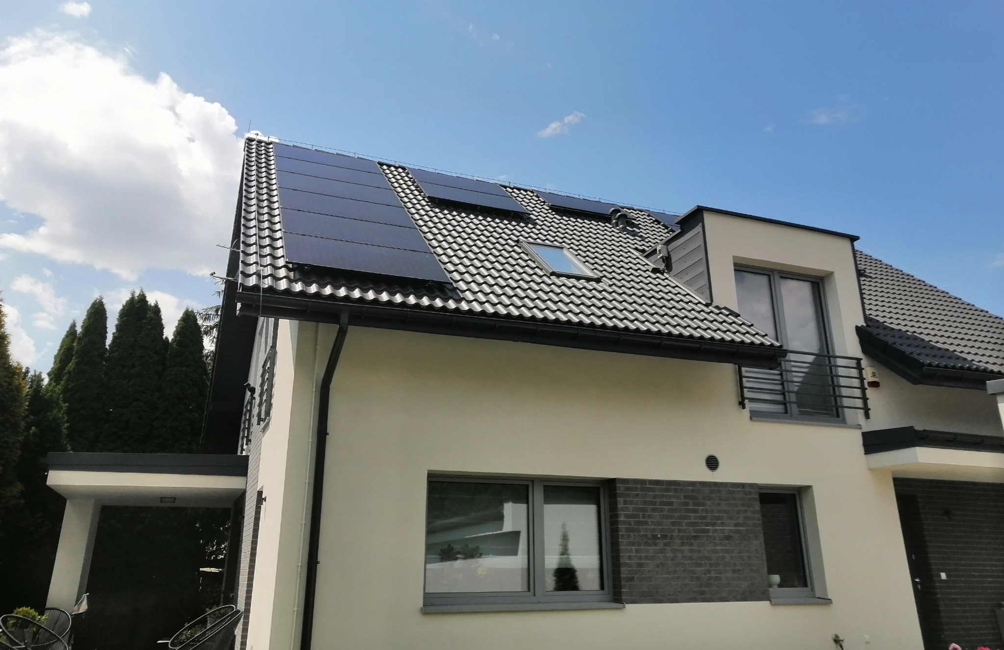 Nowoczesny dom jednorodzinny z panelami fotowoltaicznymi na dachu pokrytym szarą dachówką, widok z dołu na tle błękitnego nieba.