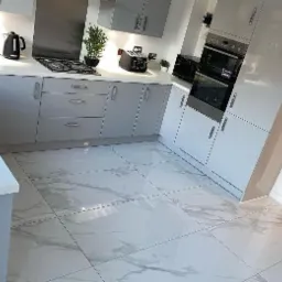 Nowoczesna kuchnia z szarymi szafkami na wysoki połysk i białymi marmurkowymi płytkami podłogowymi, widok na zabudowane piekarniki i sprzęty AGD.