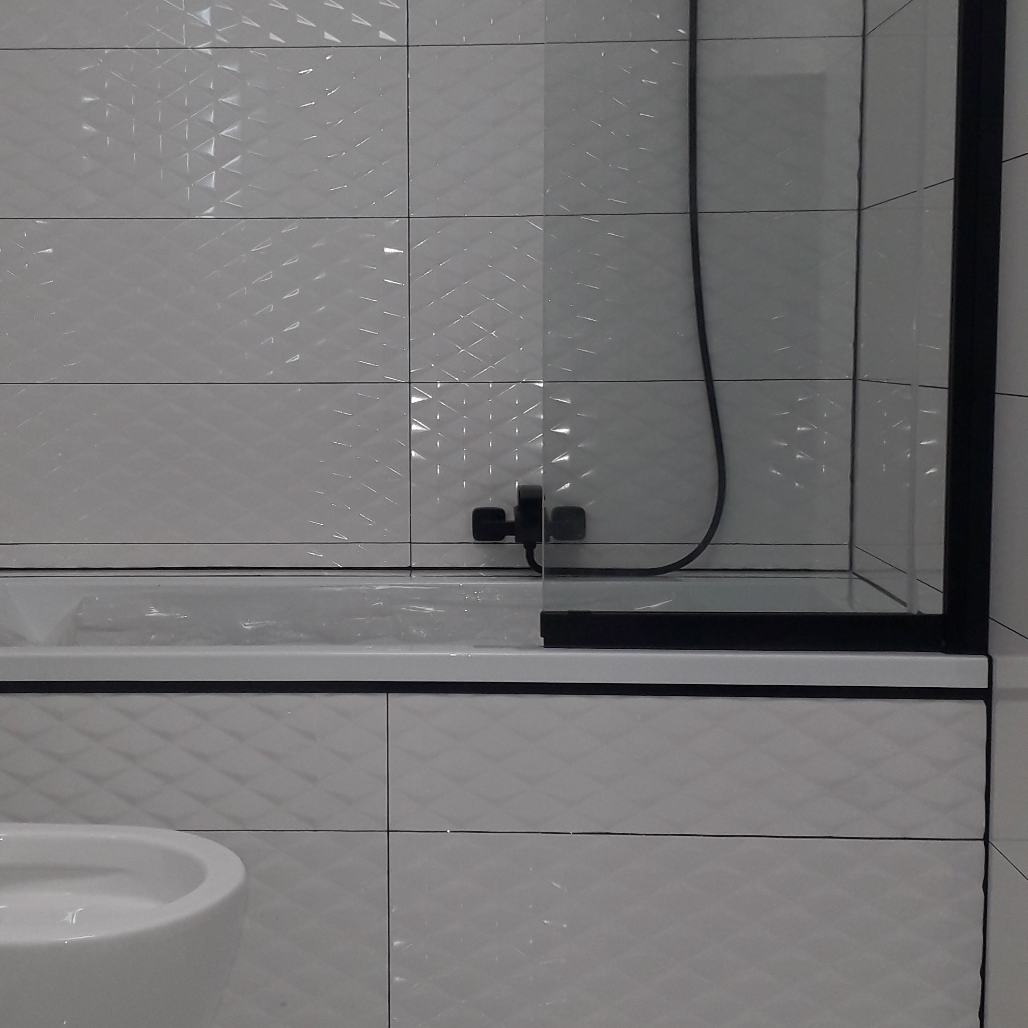 Fragment łazienki z białą, wzorzystą glazurą, czarną armaturą i szklanym parawanem nawannowym z czarną ramą, widoczny fragment toalety.