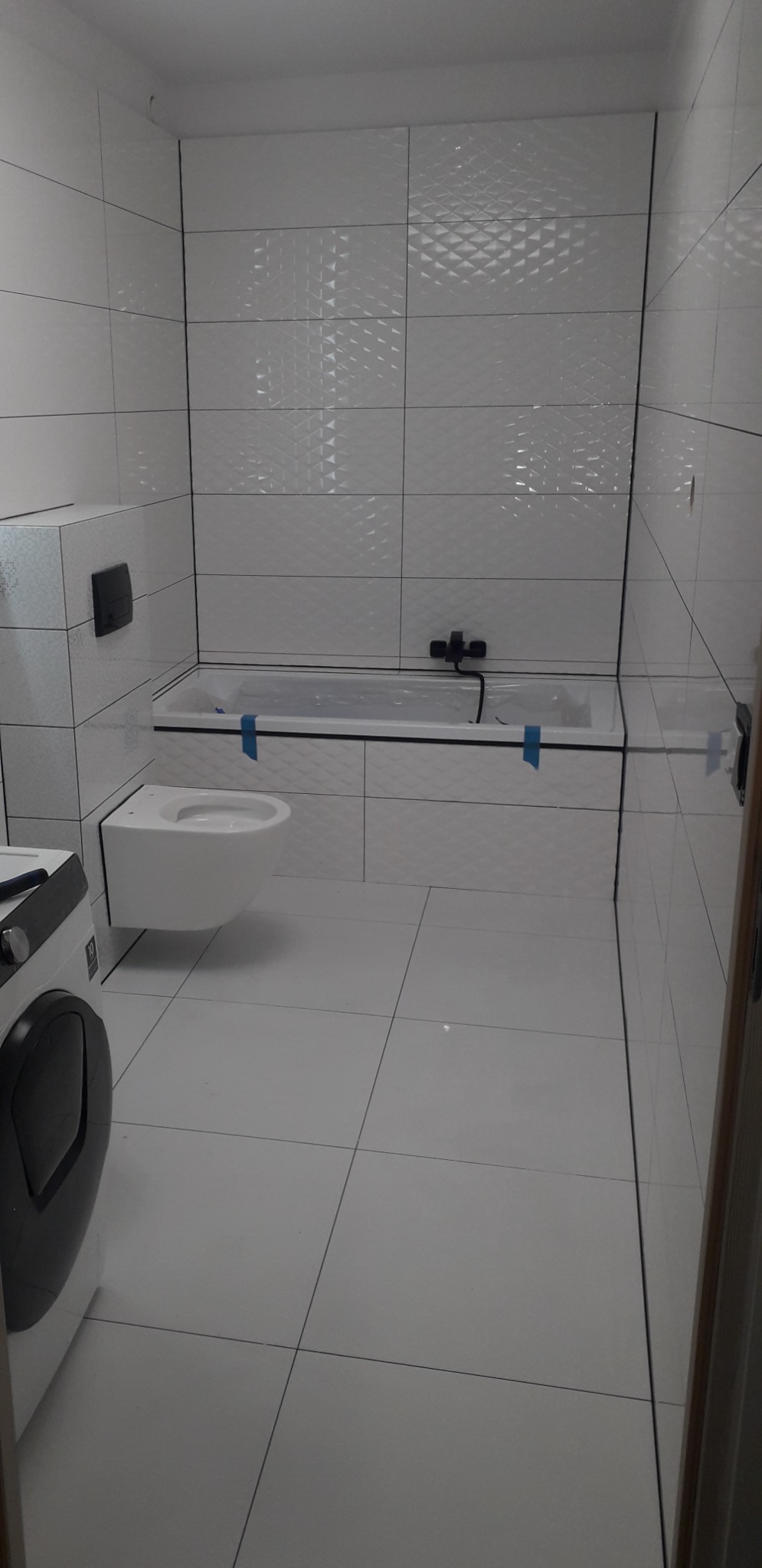 Jasna łazienka w Szczecinie po remoncie, z białą armaturą i płytkami z subtelnym wzorem, widoczna pralka i taśma malarska na wannie.