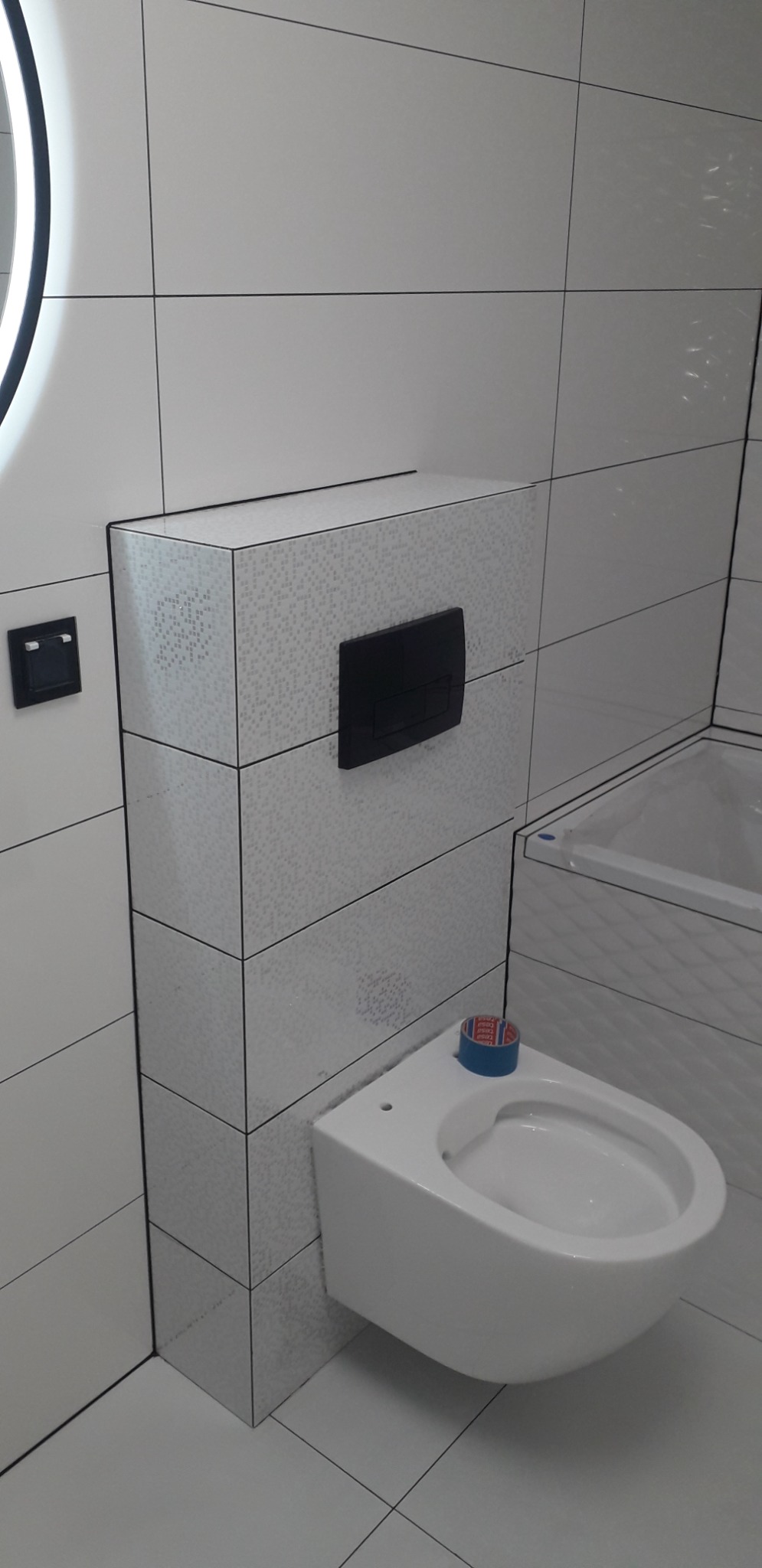 Podwieszana toaleta z czarnym przyciskiem spłukującym, obudowana płytkami imitującymi mozaikę, częściowo zabezpieczona niebieską taśmą malarską, na tle białych płytek ściennych i fragmentu wanny.