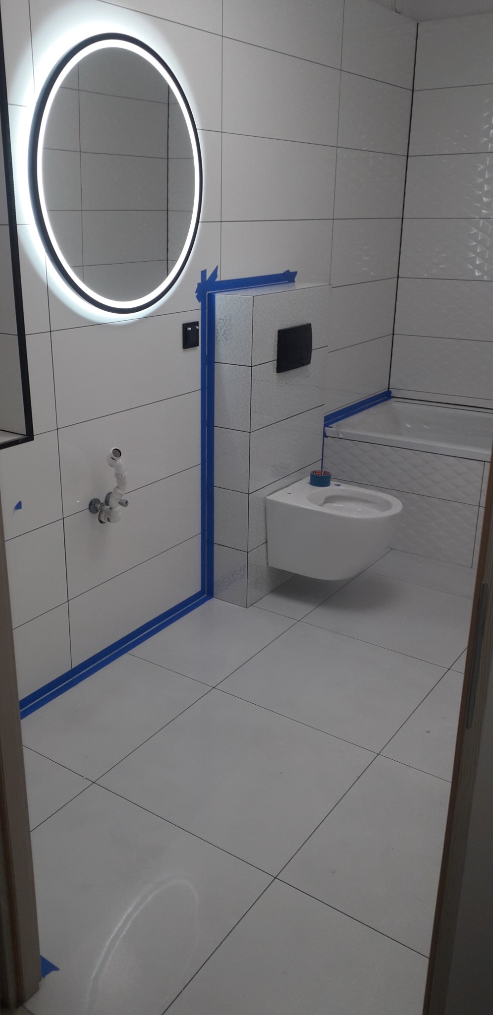 Biała łazienka w trakcie prac wykończeniowych, widoczne płytki, podwieszana toaleta, okrągłe lustro z podświetleniem LED oraz niebieska taśma malarska zabezpieczająca krawędzie.