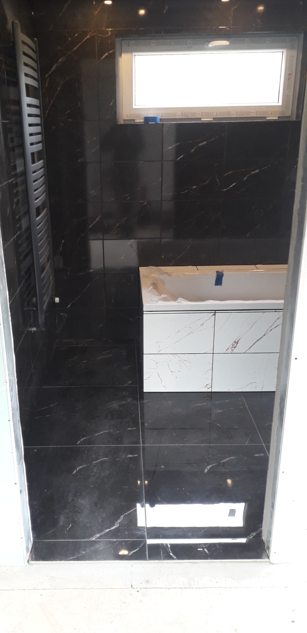 Częściowo wykończona łazienka z czarnymi płytkami imitującymi marmur na ścianach i podłodze, biała obudowa wanny z panelami imitującymi marmur, grzejnik drabinkowy i okno z białą ramą.