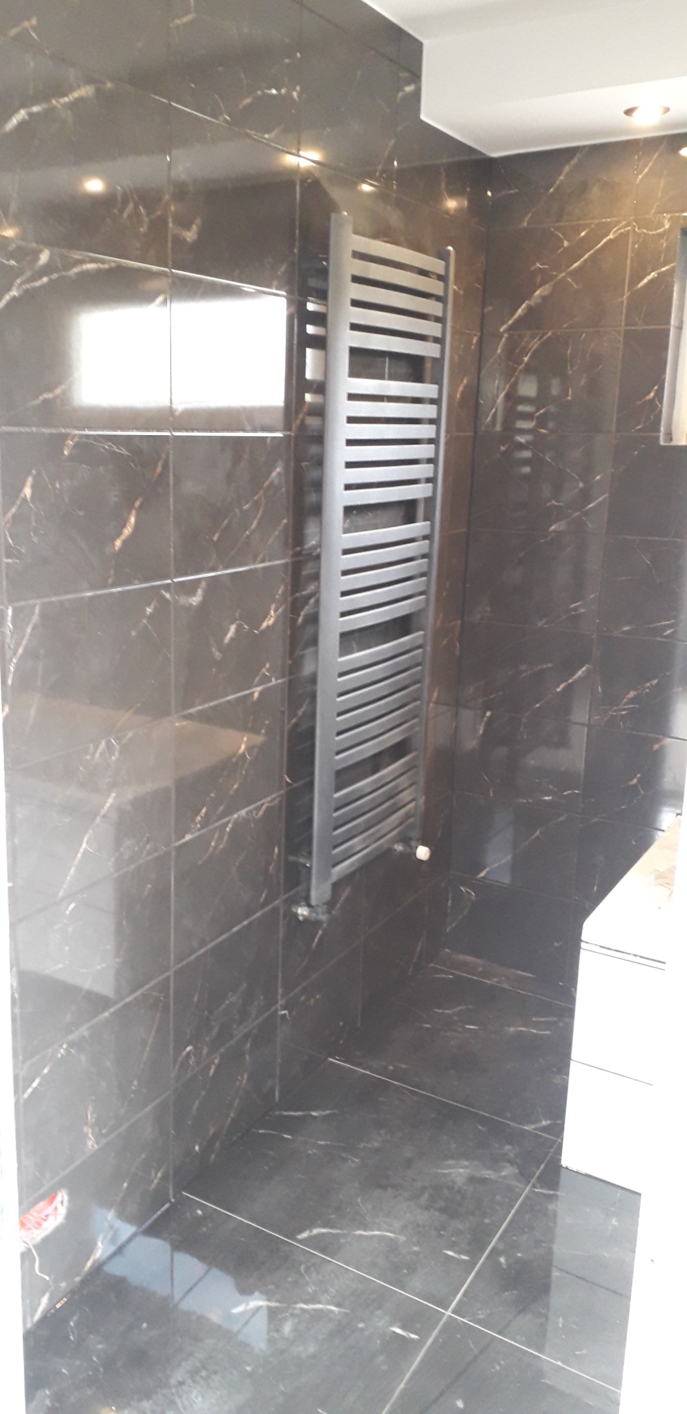 Nowoczesny, srebrny grzejnik drabinkowy zamontowany na ścianie z ciemnych, marmurkowych płytek w łazience. Widoczny fragment podłogi z identycznych płytek i narożnik ściany.