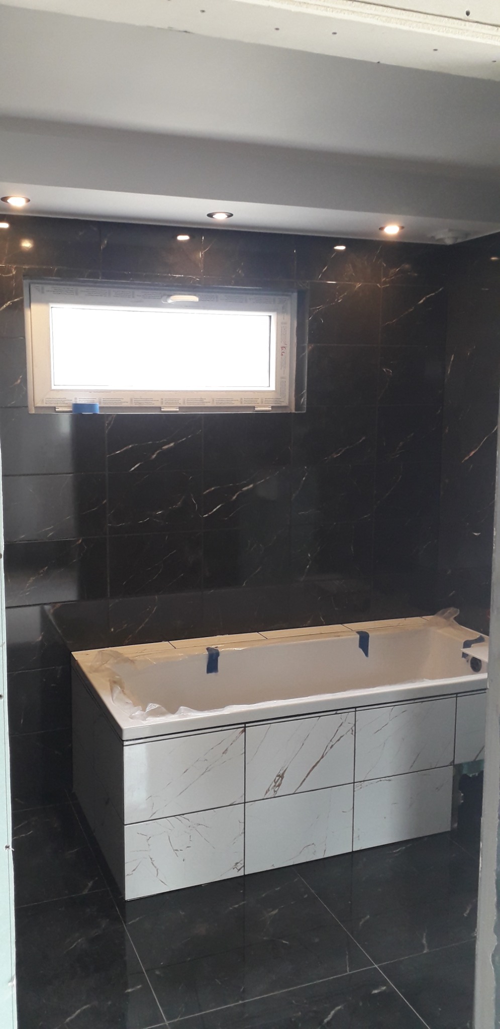 Wanna obudowana białymi płytkami imitującymi marmur, zabezpieczona folią i taśmą malarską, na tle ściany wyłożonej ciemnymi płytkami z marmurowym wzorem, małe okno w górnej części ściany...