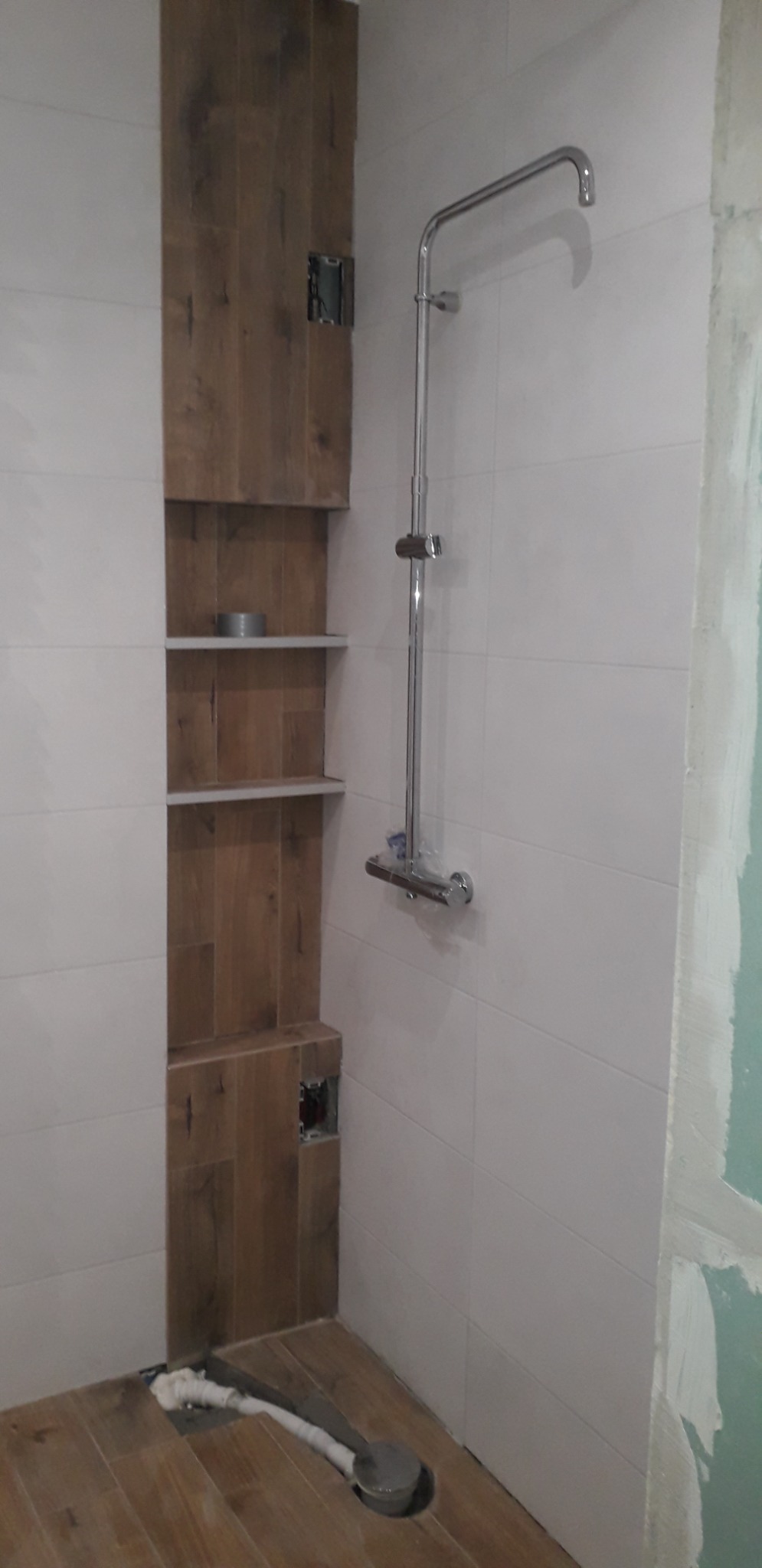 Wnęka z półkami wyłożona płytkami imitującymi drewno, zamontowany panel prysznicowy w trakcie remontu łazienki, widoczne niedokończone prace wykończeniowe.