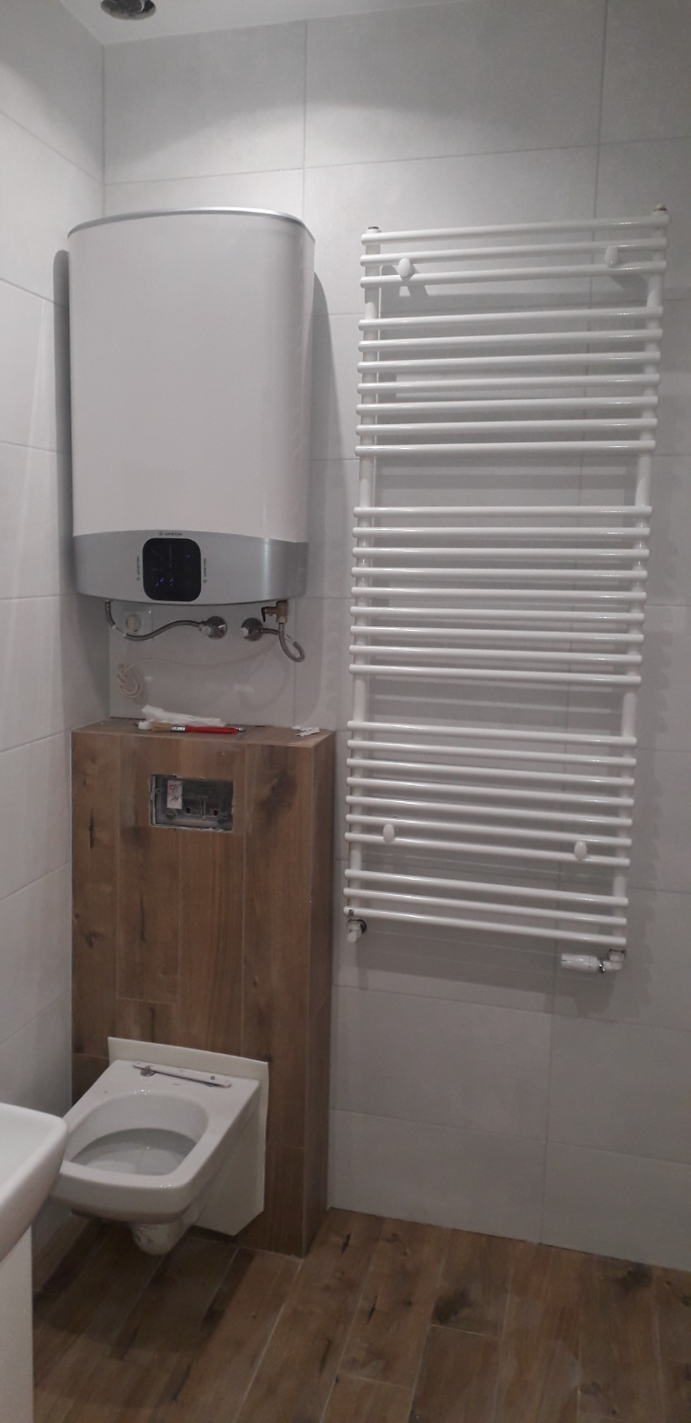 Świeżo wykończona łazienka z podwieszaną toaletą, bojlerem i grzejnikiem drabinkowym na tle białych płytek i drewnopodobnej podłogi.