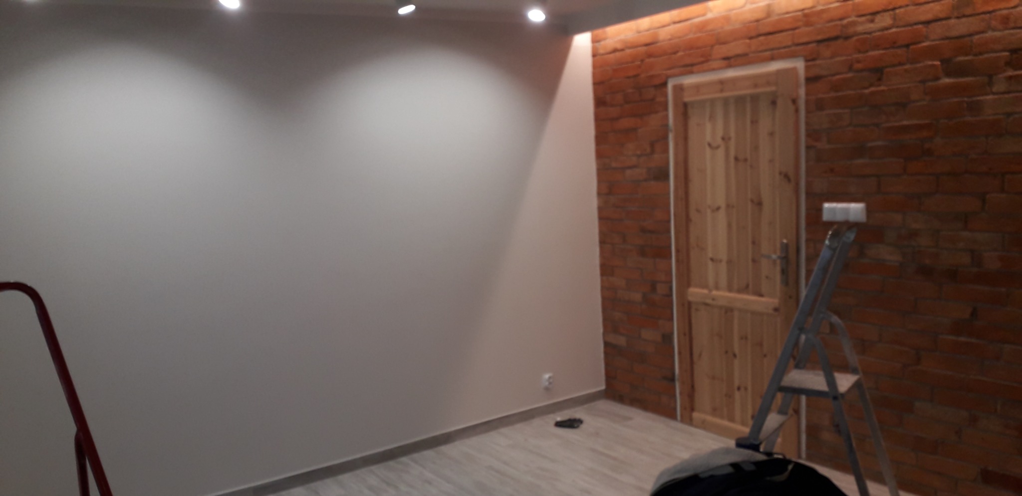 Świeżo pomalowane, jasnoszare ściany w pokoju z ceglaną ścianą i drewnianymi drzwiami, widoczna drabina malarska i fragment drugiej drabiny.