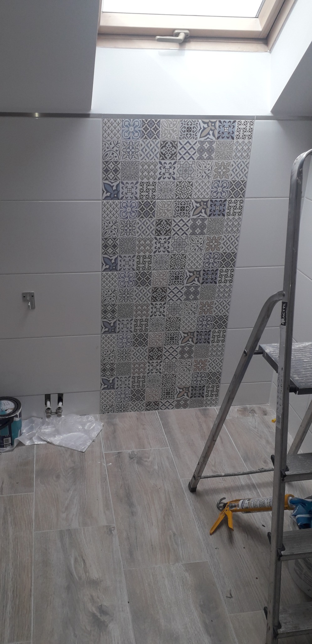 Fragment łazienki w trakcie prac wykończeniowych; widoczny fragment ściany z ułożonymi płytkami w geometryczne wzory, drabina aluminiowa, puszka z farbą i pistolet do silikonu na podłodze...
