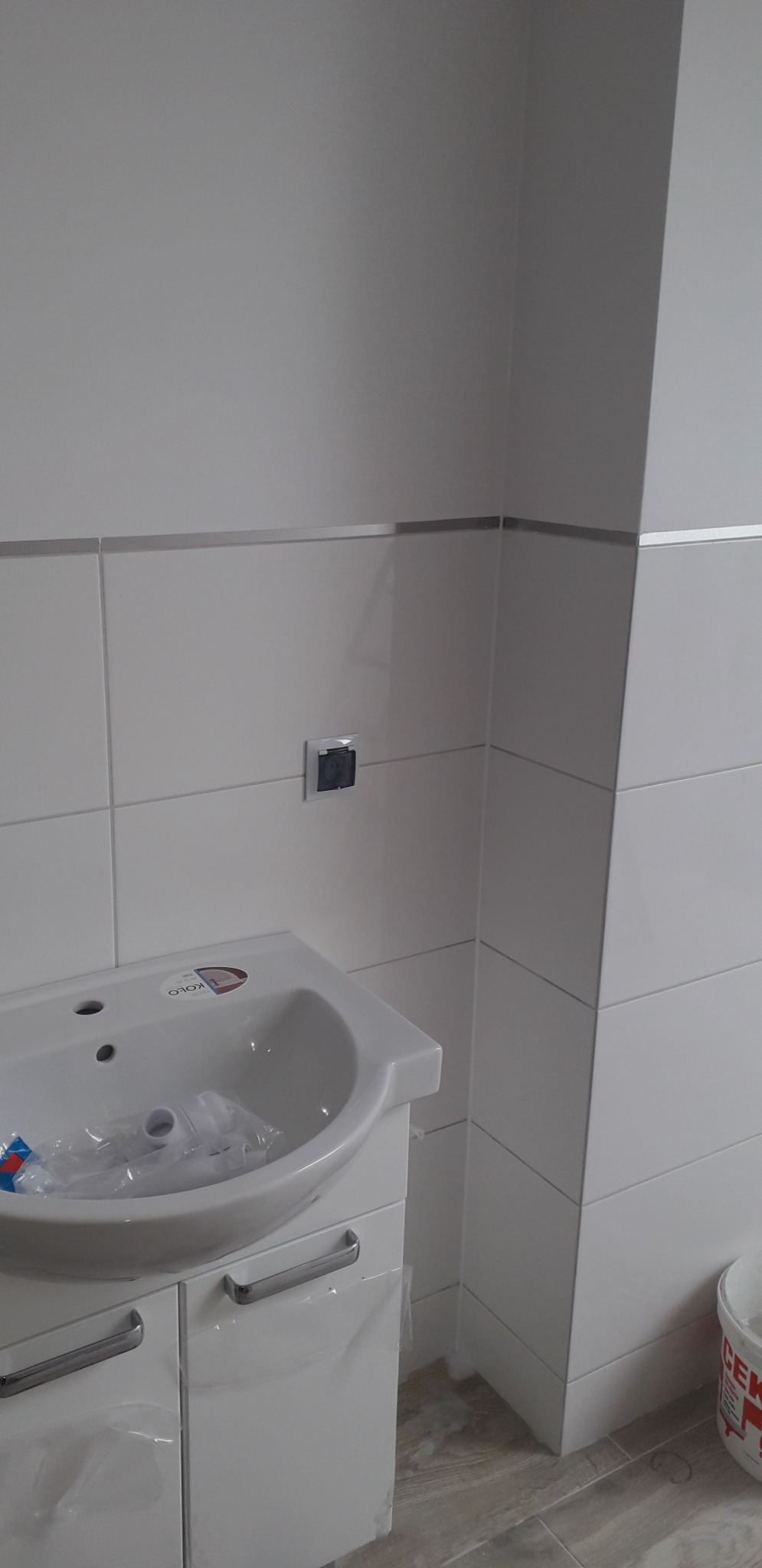 Biała umywalka z szafką w trakcie montażu w łazience z jasnymi płytkami, widoczna niezapakowana armatura i wiadro z zaprawą.