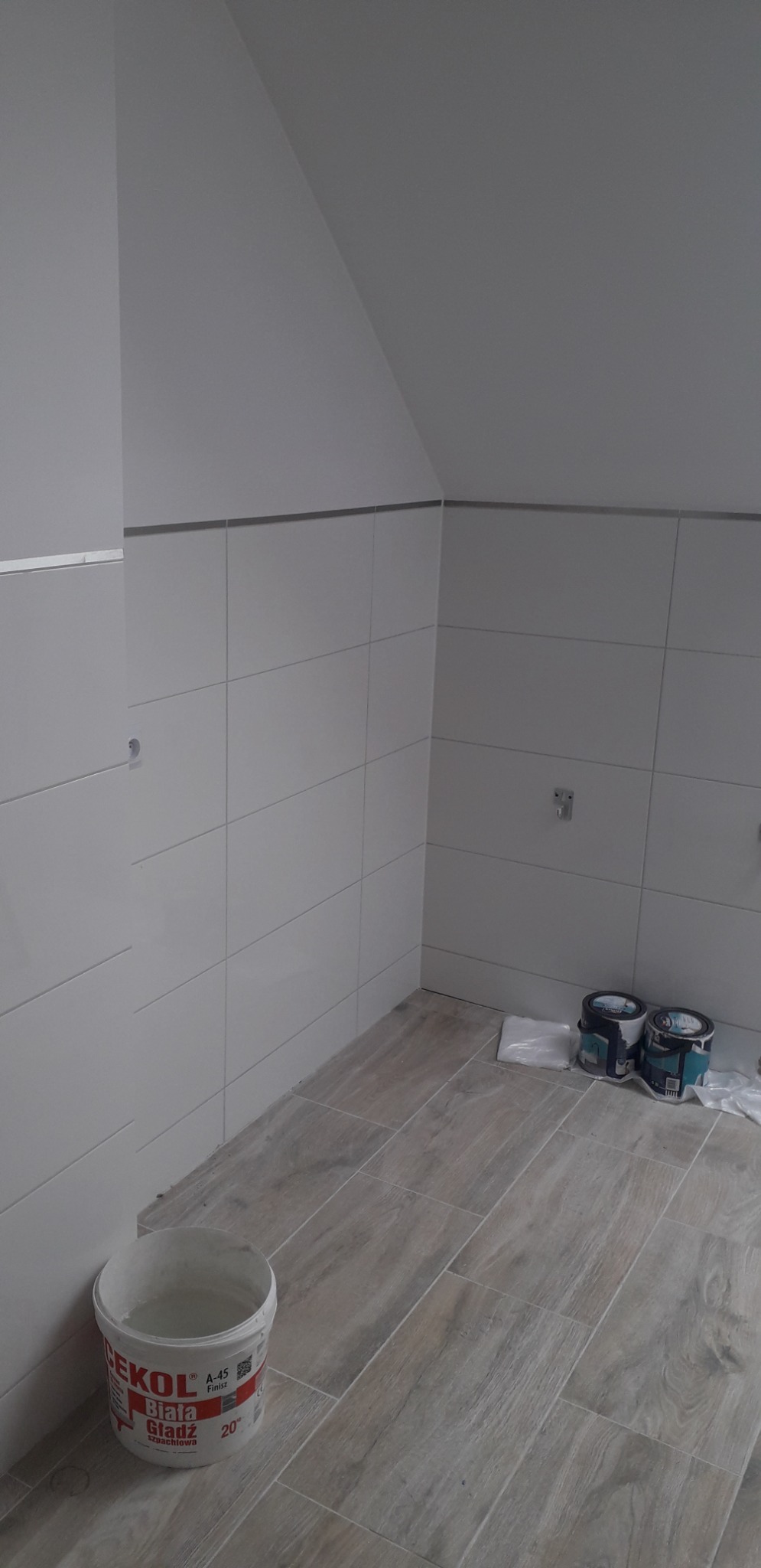 Wnętrze łazienki w trakcie prac wykończeniowych: białe płytki ścienne z metalową listwą, podłoga wyłożona płytkami imitującymi drewno, wiadro z gładzią szpachlową i puszki z farbą na podłodze...