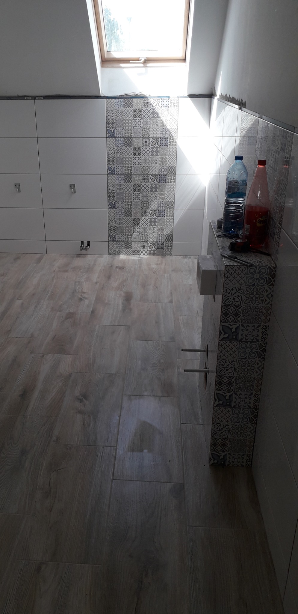 Wnętrze łazienki w trakcie prac: ułożona podłoga imitująca drewno, częściowo wykończone ściany z białymi płytkami i dekoracyjnym pasem płytek w geometryczne wzory, widoczne instalacje, okno dachowe...