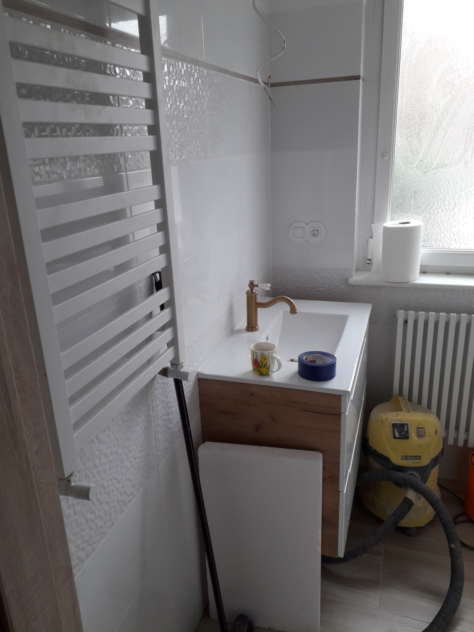 Remont łazienki w trakcie: Umywalka z drewnianą szafką, złota bateria, kubek w kwiaty, niebieska taśma malarska, białe płytki z delikatnym wzorem, grzejnik drabinkowy, odkurzacz przemysłowy Karcher...