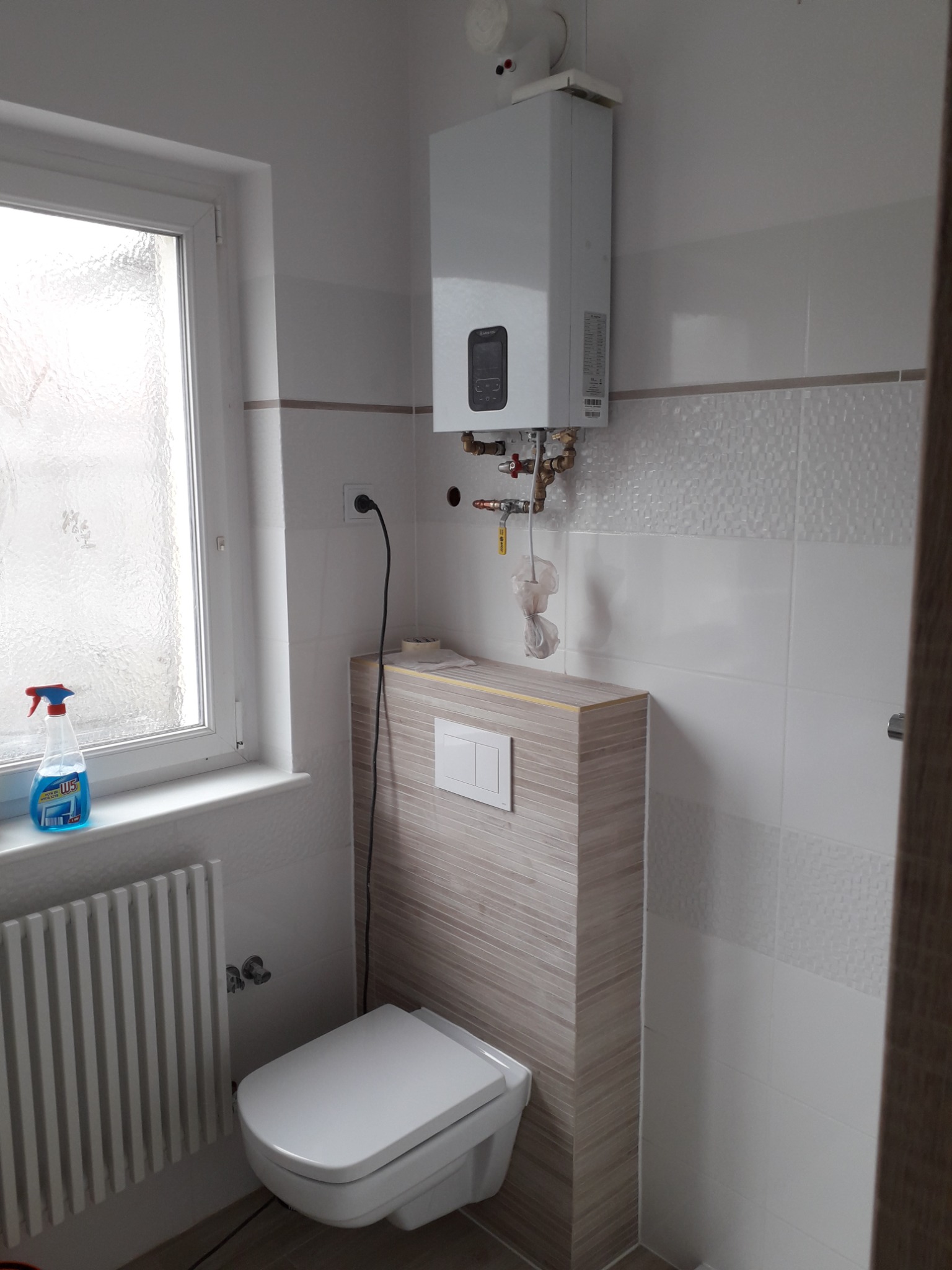 Nowoczesna łazienka z podwieszaną toaletą, grzejnikiem żeberkowym, bojlerem elektrycznym i oknem z matową szybą. Na parapecie butelka płynu do mycia okien.