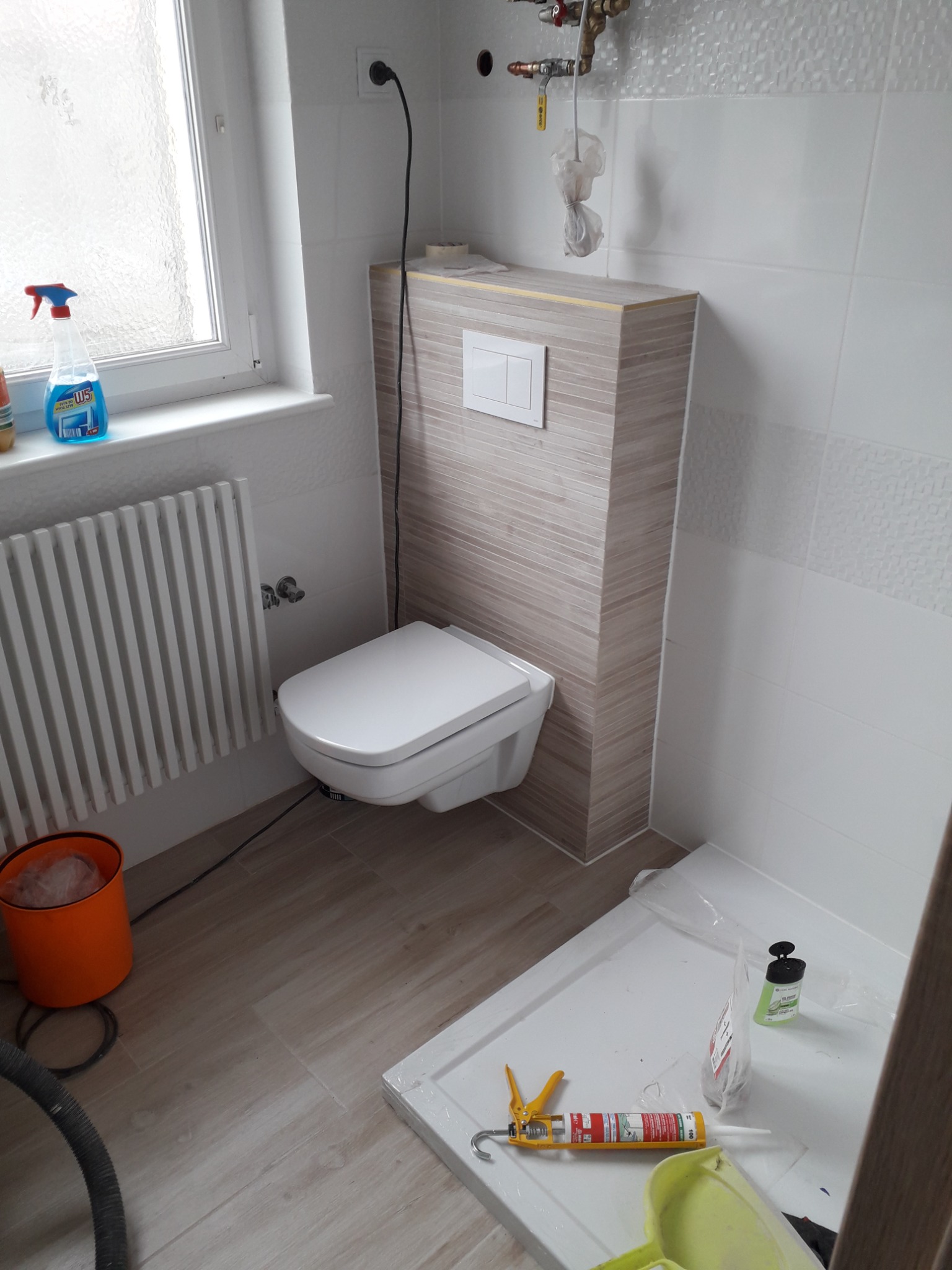 Podwieszana toaleta z zabudową w łazience w trakcie prac wykończeniowych, widoczna kabina prysznicowa w folii, narzędzia i chemia budowlana.