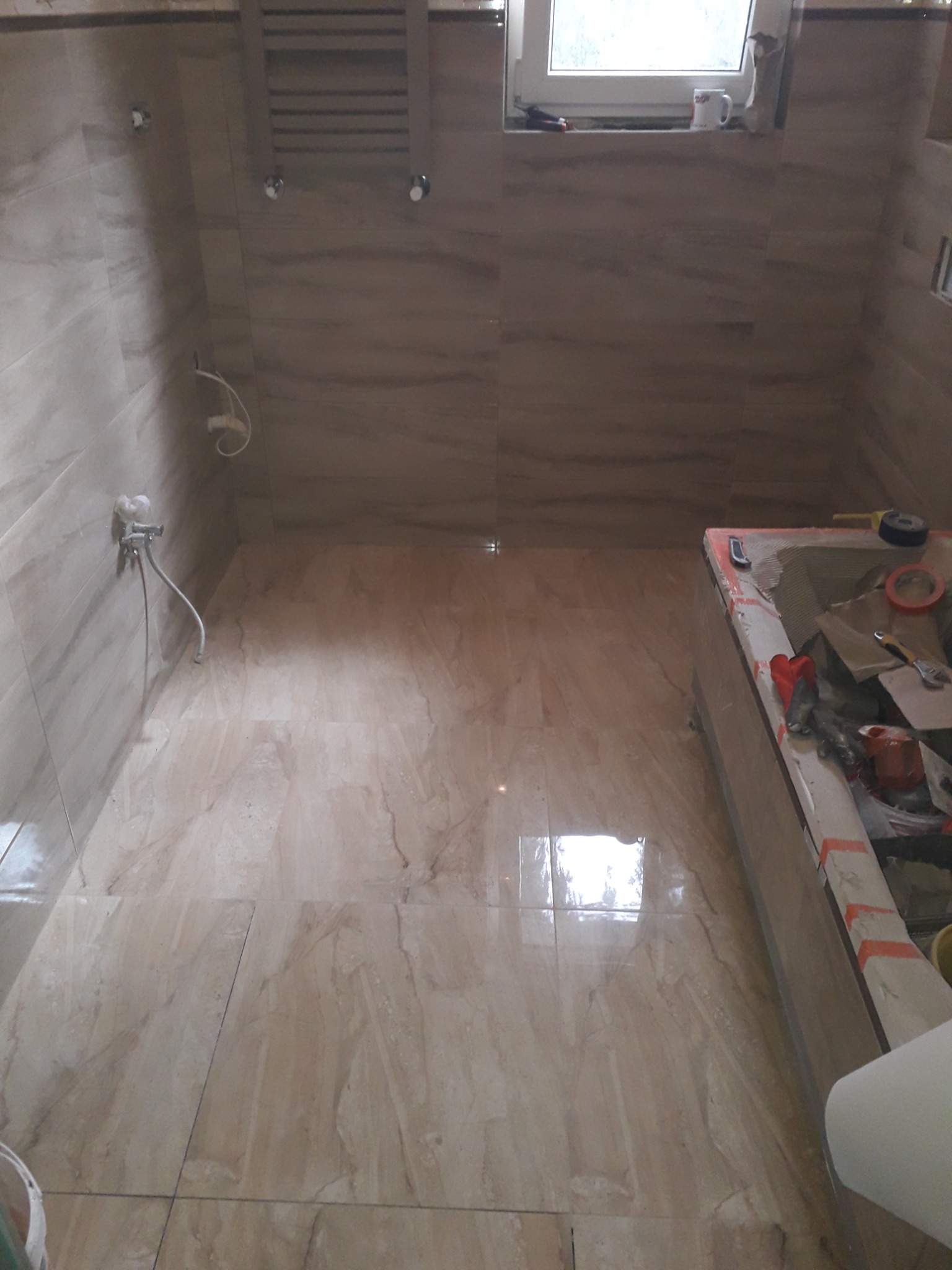 Łazienka w trakcie prac wykończeniowych, widoczne płytki imitujące marmur na ścianach i podłodze, narzędzia i materiały budowlane przy wannie, grzejnik drabinkowy na ścianie.