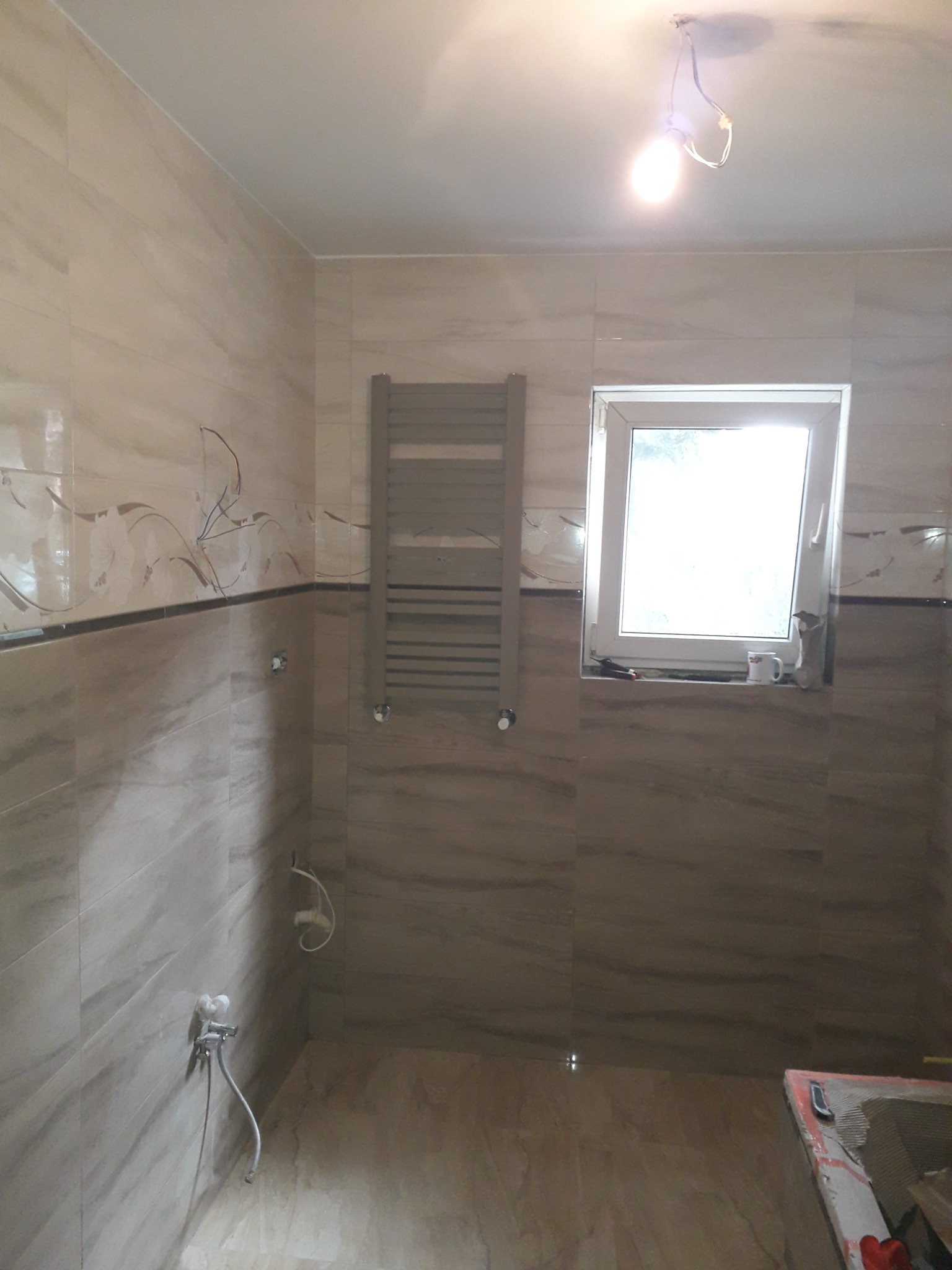 Wnętrze łazienki w trakcie prac wykończeniowych: płytki imitujące marmur na ścianach i podłodze, grzejnik drabinkowy, okno z kubkiem na parapecie, widoczne przewody elektryczne i niedokończone...