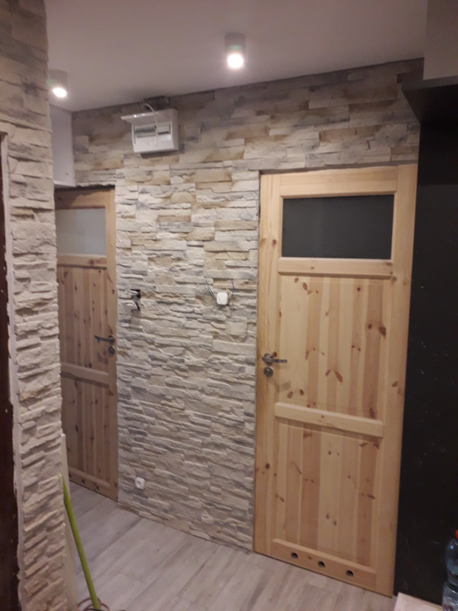Wykończone wnętrze przedpokoju z dwiema drewnianymi drzwiami, ściany pokryte dekoracyjnym kamieniem w odcieniach beżu i brązu, podłoga wyłożona panelami imitującymi drewno, widoczna instalacja...