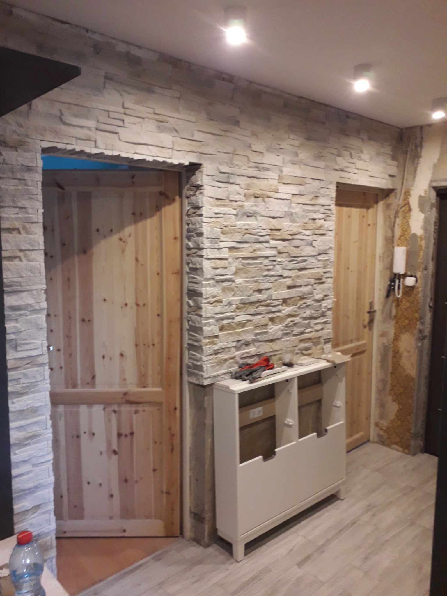 Ściana wykończona dekoracyjnym kamieniem, drewniane drzwi z surowego drewna, biała szafka z narzędziami i rękawicami roboczymi na blacie, podłoga z płytek imitujących drewno, widoczne ślady po...