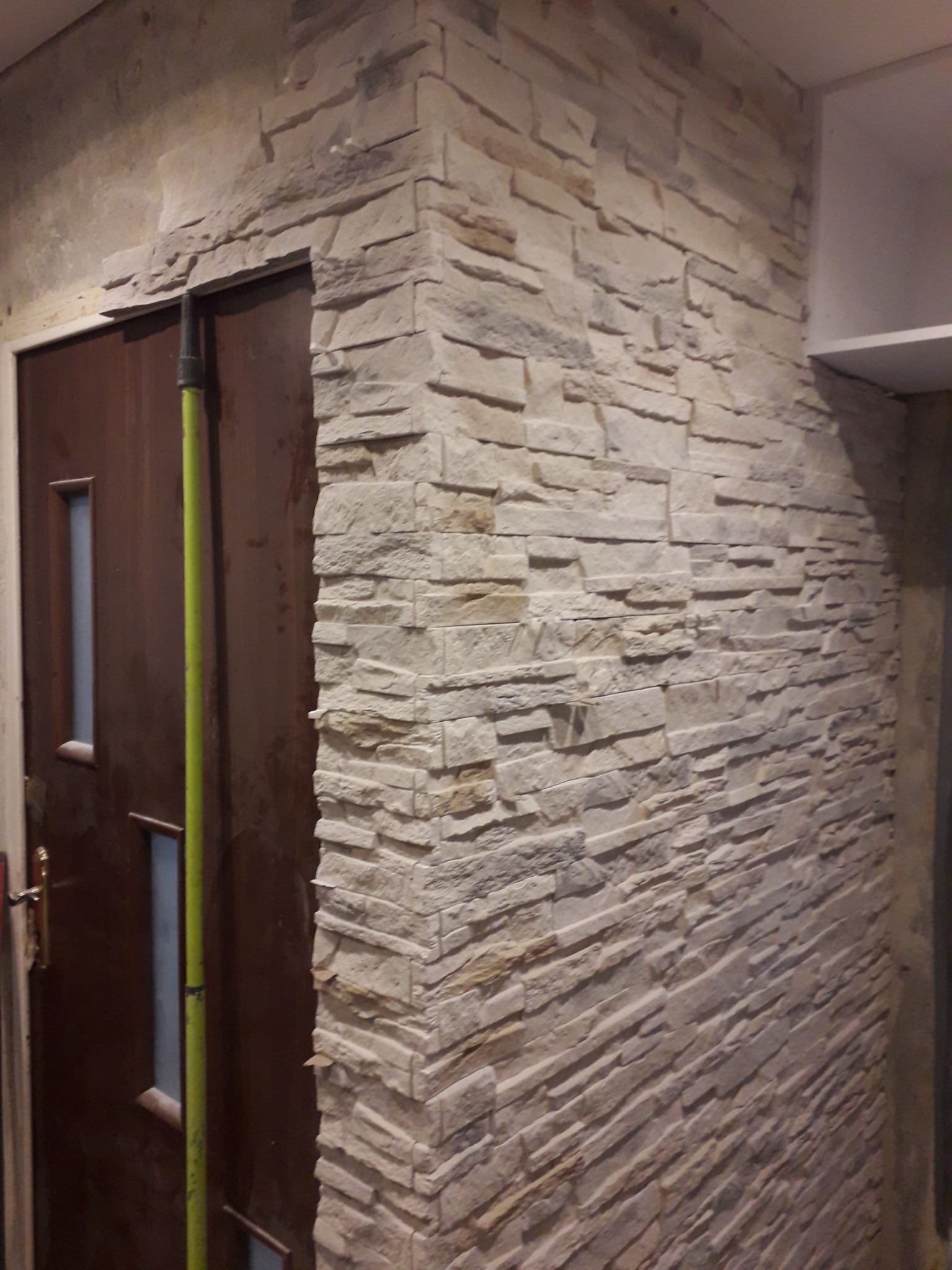 Fragment ściany wykończonej dekoracyjnym kamieniem elewacyjnym w odcieniach beżu i szarości, przylegający do ościeżnicy brązowych drzwi z przeszkleniem, widoczna zielona poziomica oparta o drzwi.